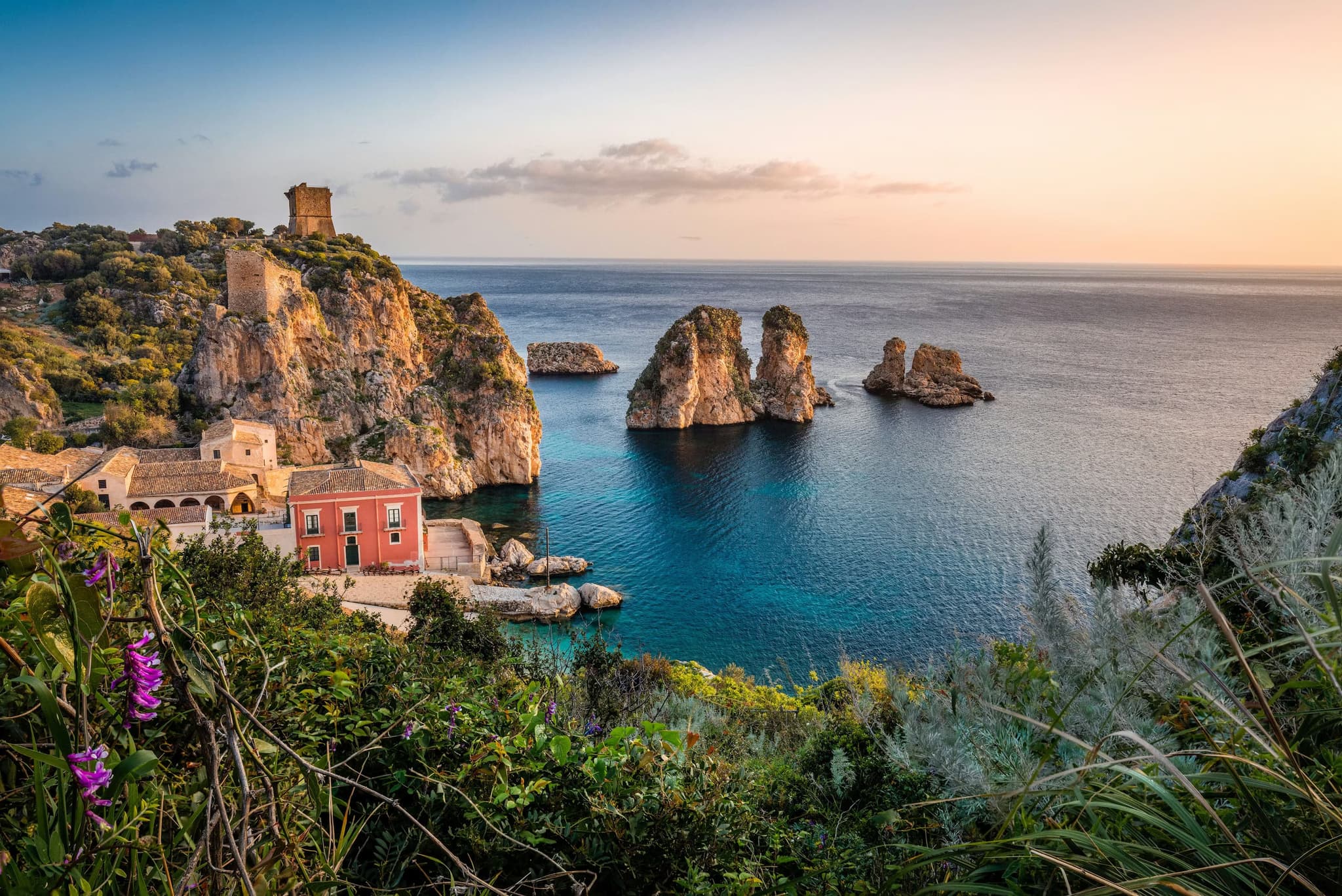 Sunset view of a coastal village with rocky cliffs and sea stacks, a historic watchtower, and pink flowers in the foreground, exuding tranquility.