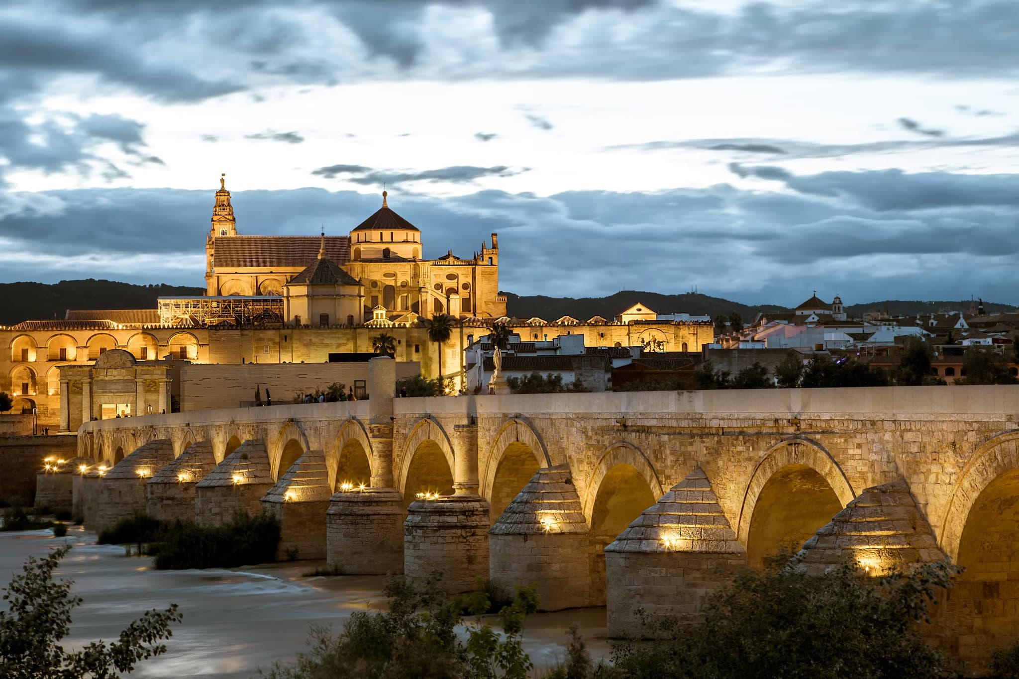 Illuminated Roman bridge leading to the historic mosque-cathedral in Córdoba, Spain, at twilight. The scene exudes a serene, historic ambiance.