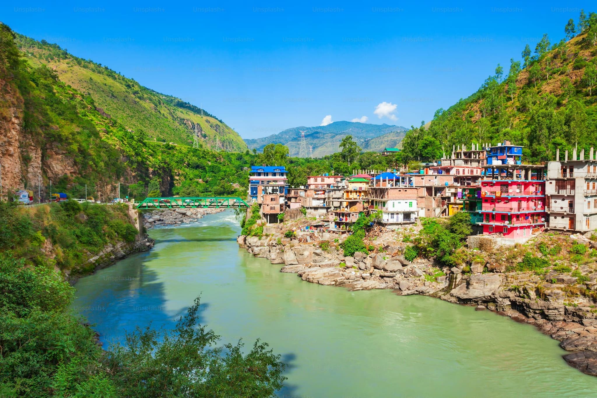 Colorful houses line a rocky riverbank, surrounded by lush green hills under a blue sky. A bridge spans the river, creating a scenic, vibrant view.