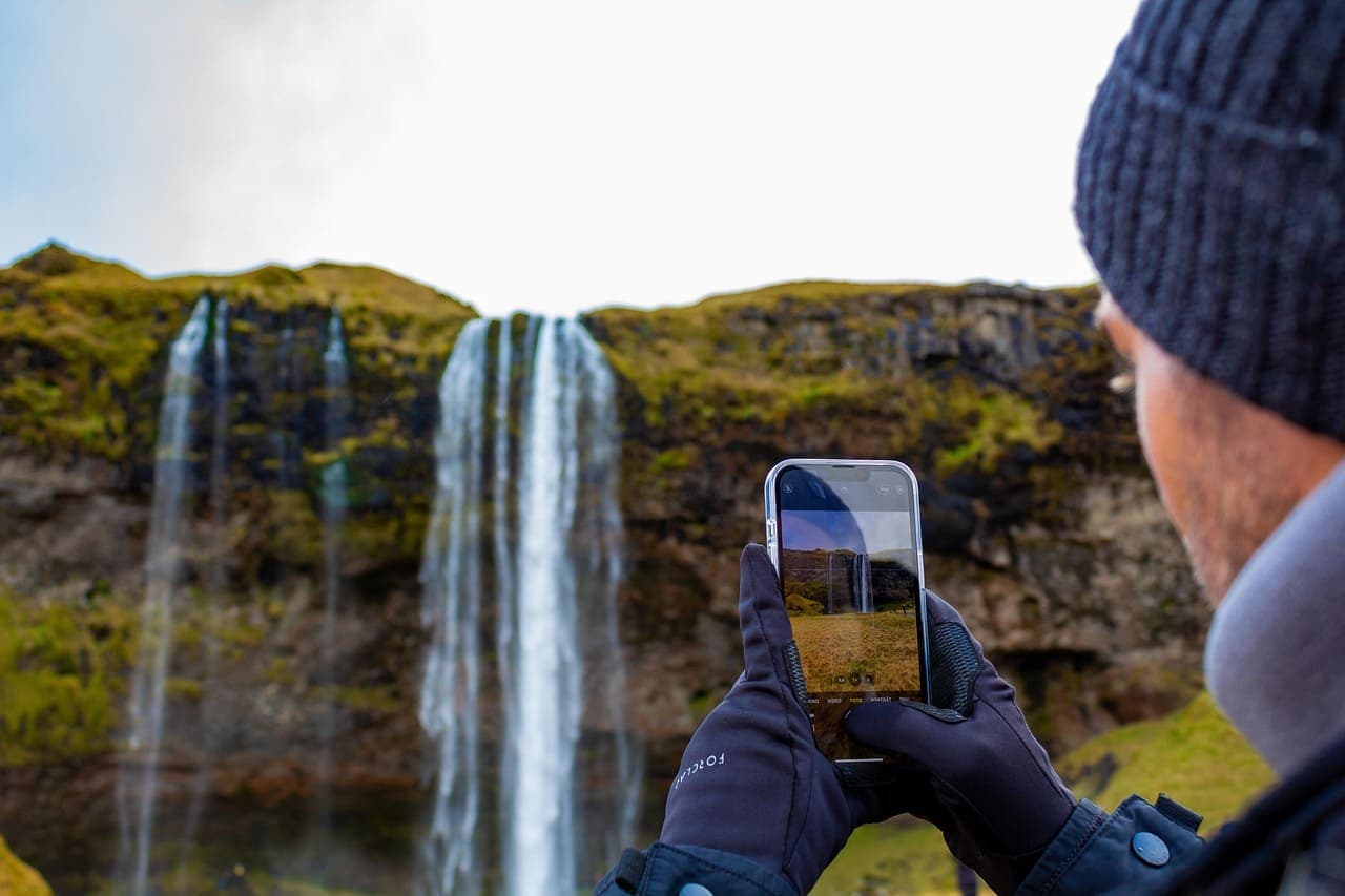 Traveller photographing a waterfall in Iceland with a smartphone, standing near cascading water and moss covered cliffs.