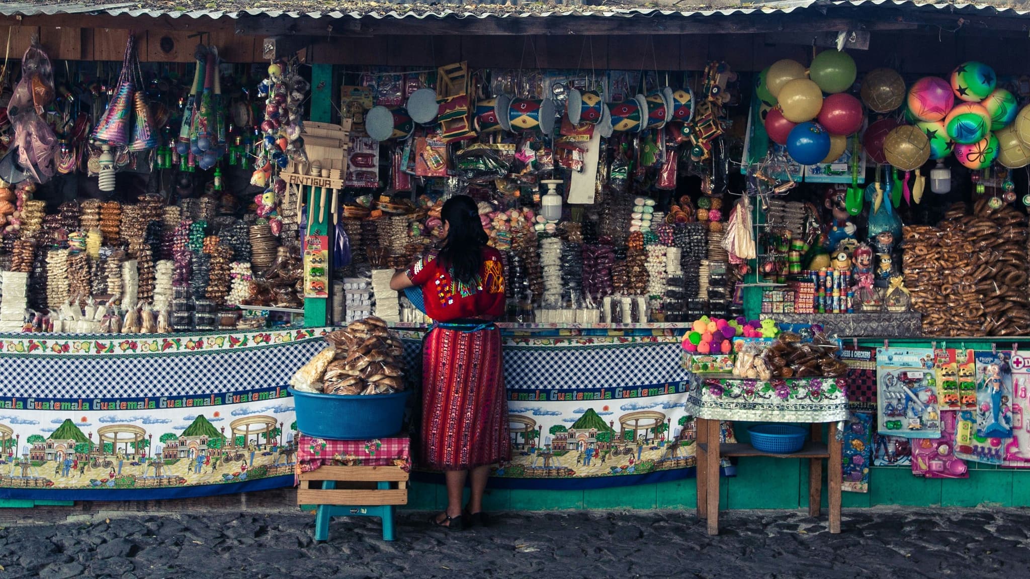 Local market stall in Guatemala with colourful crafts and snacks, as a woman in traditional clothing stands in front of the display.