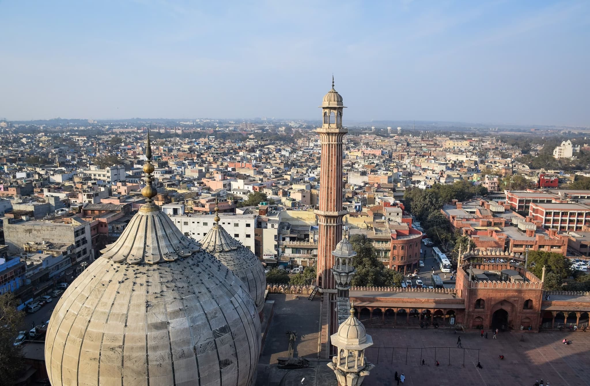 Aerial view of a bustling cityscape featuring a historic mosque with large domes and a tall minaret in the foreground. The scene is vibrant and expansive.