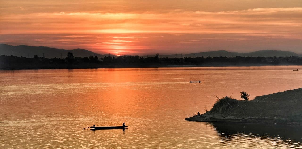 Peaceful sunset over the Mekong River with a small boat floating on the water and warm orange light reflecting across the surface.
