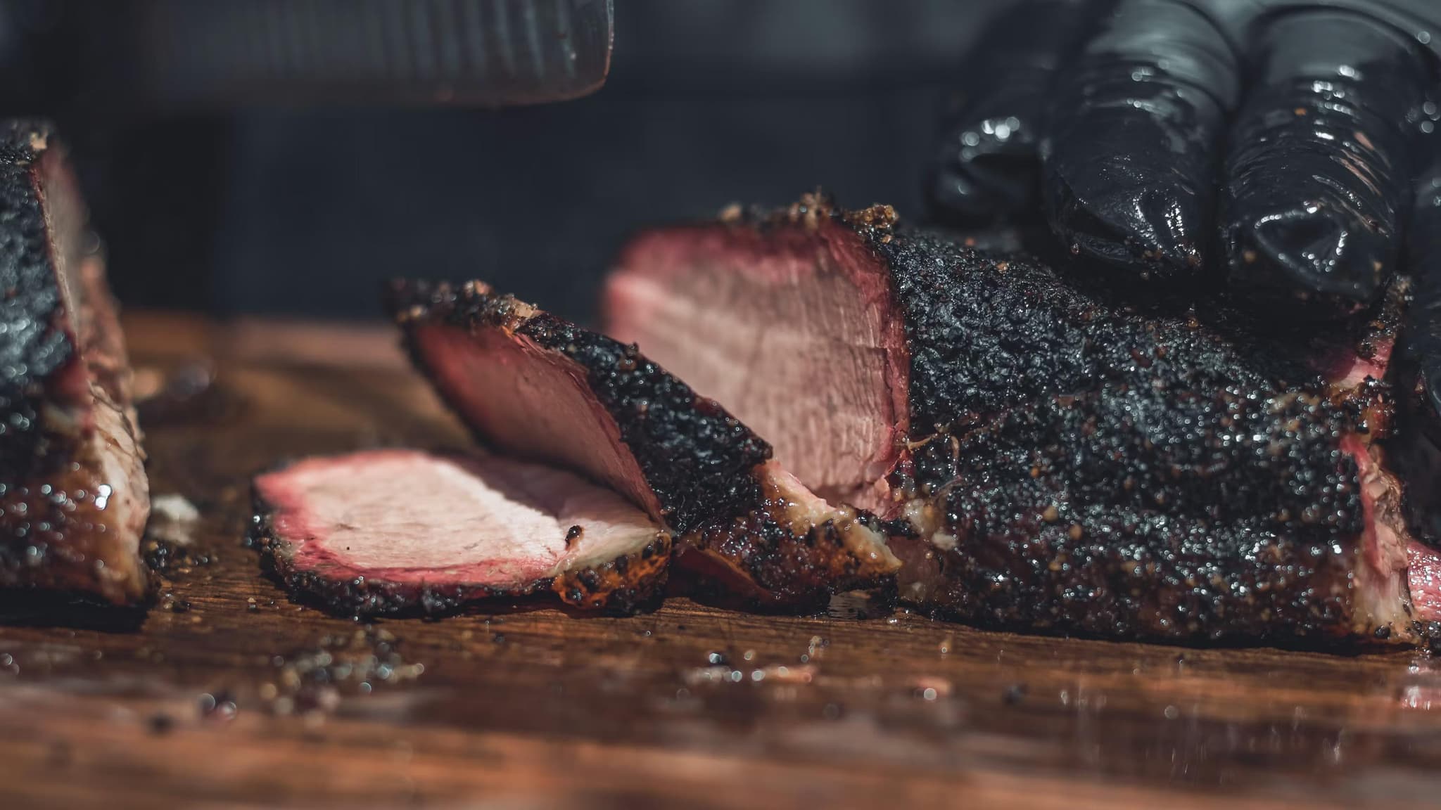 Close-up of sliced, juicy smoked meat with a blackened crust, on a wooden cutting board. A gloved hand holds the meat, creating a savory and rustic tone.
