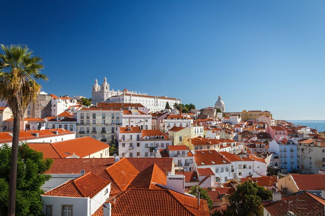 View over Lisbon’s red rooftops with white buildings and a river in the distance under a clear blue sky.