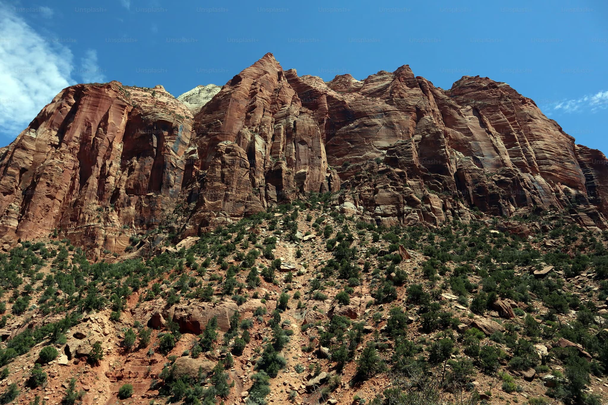 Towering red rock cliffs under a clear blue sky at Zion National Park. Sparse green vegetation dots the rugged mountain landscape. Majestic and serene.