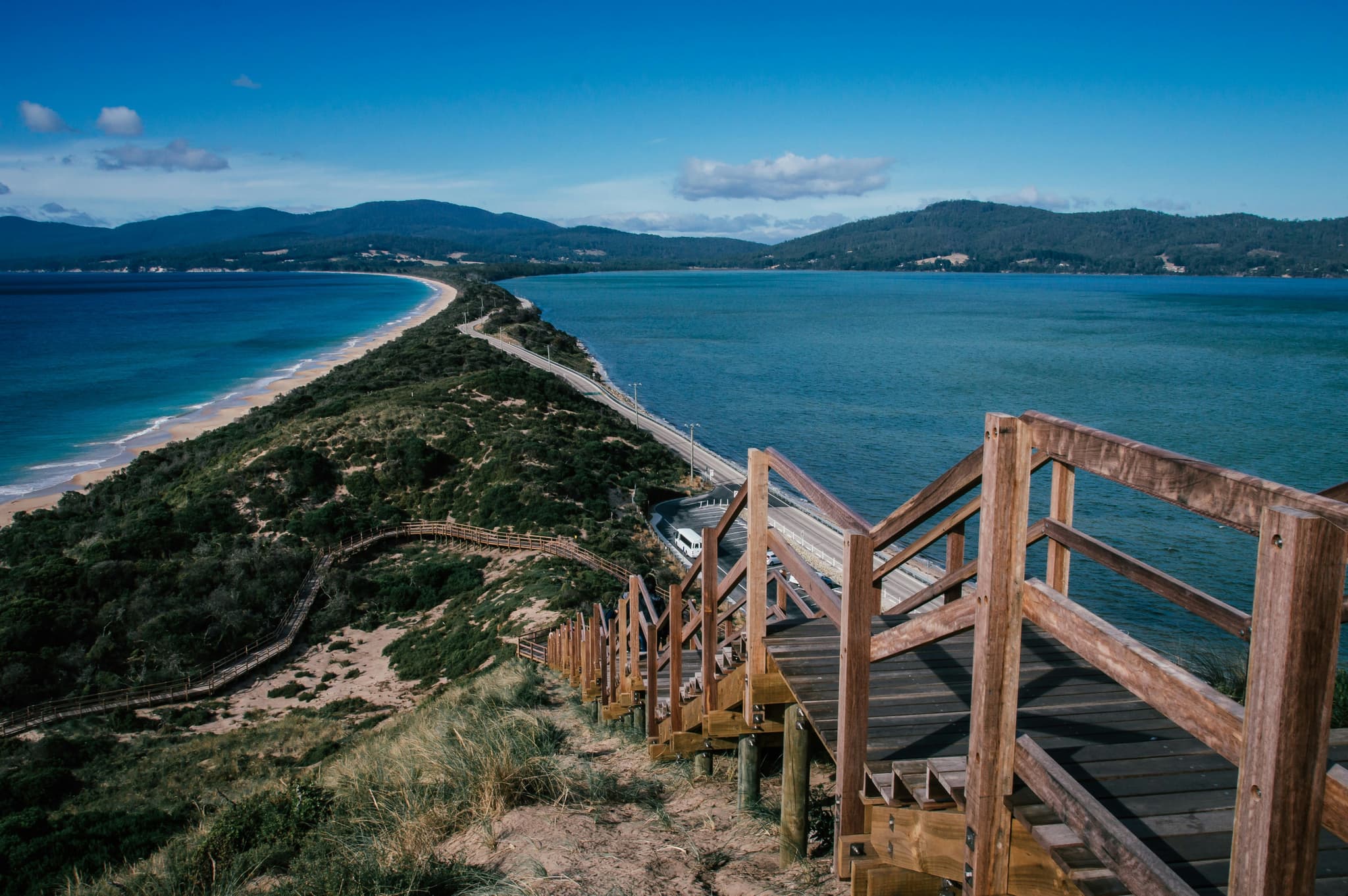 A wooden staircase on an elevated hill on the beach of Tasmania, clear blue water and mountains in the background are visible