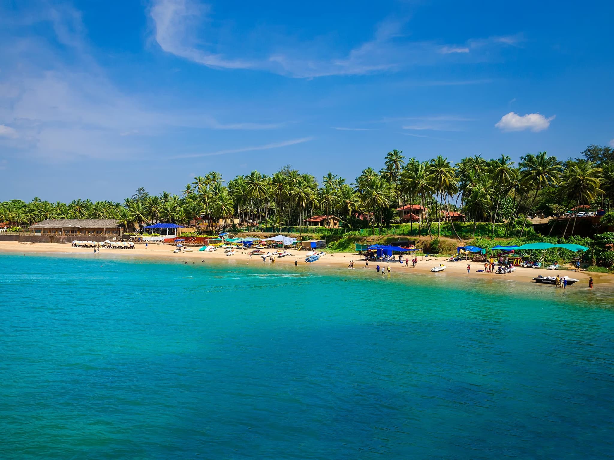 A sunlit tropical beach with turquoise water, bordered by lush palm trees and small huts. People enjoy the sand and sea under a clear blue sky.