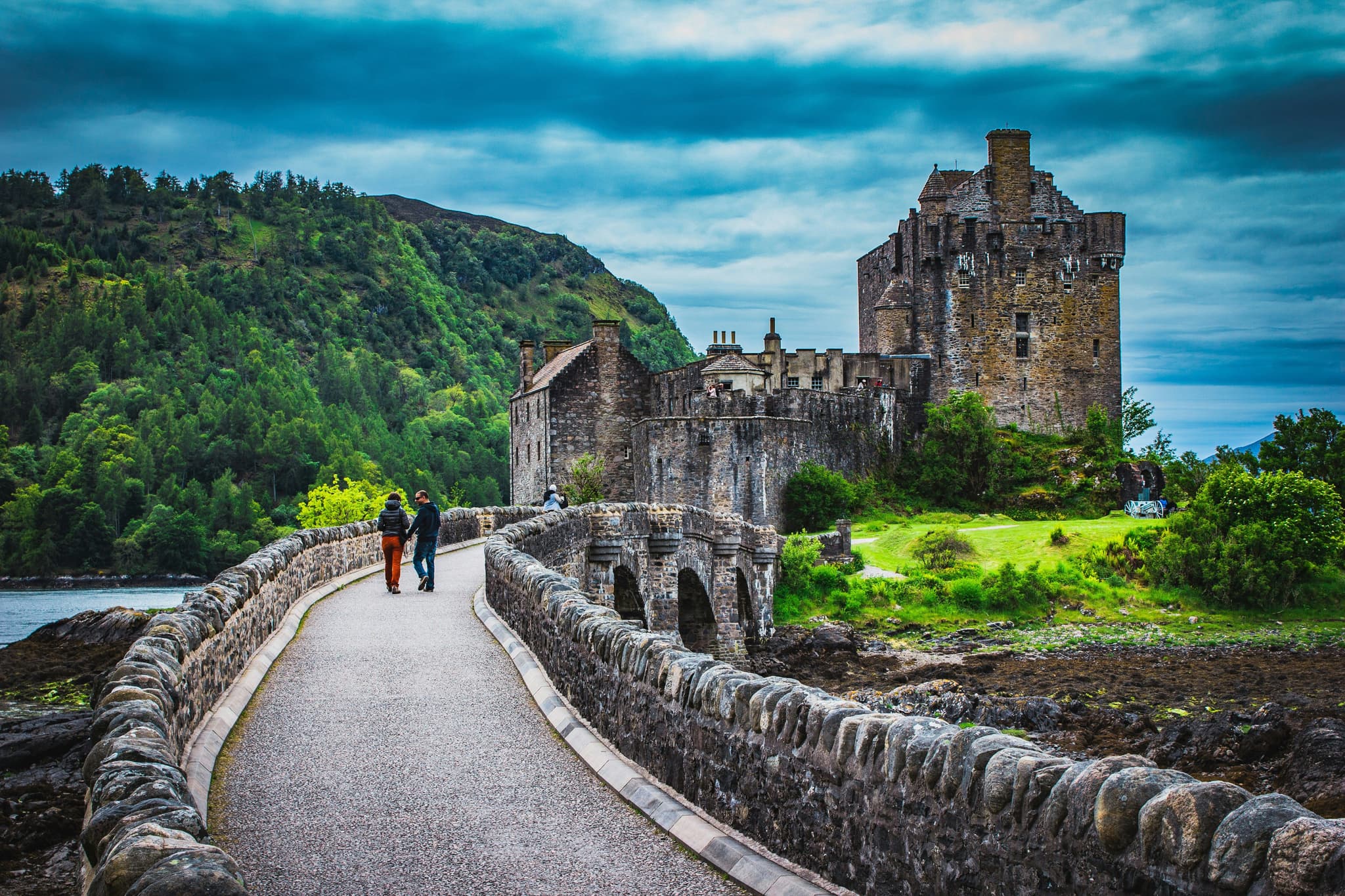 Stone bridge leading to Eilean Donan Castle surrounded by mounatins and dark blue sky