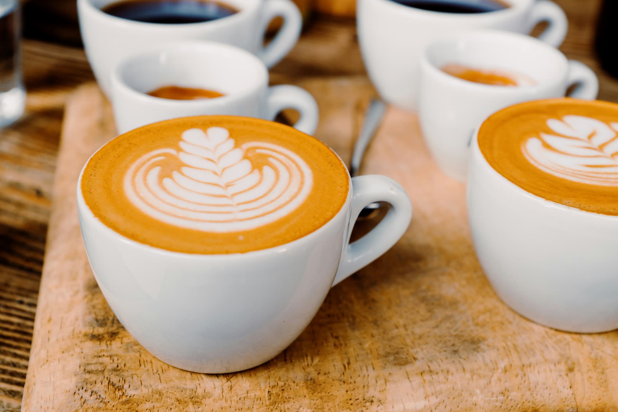 Two rows of cups of flat white coffee with a design in the foam on a wooden tray