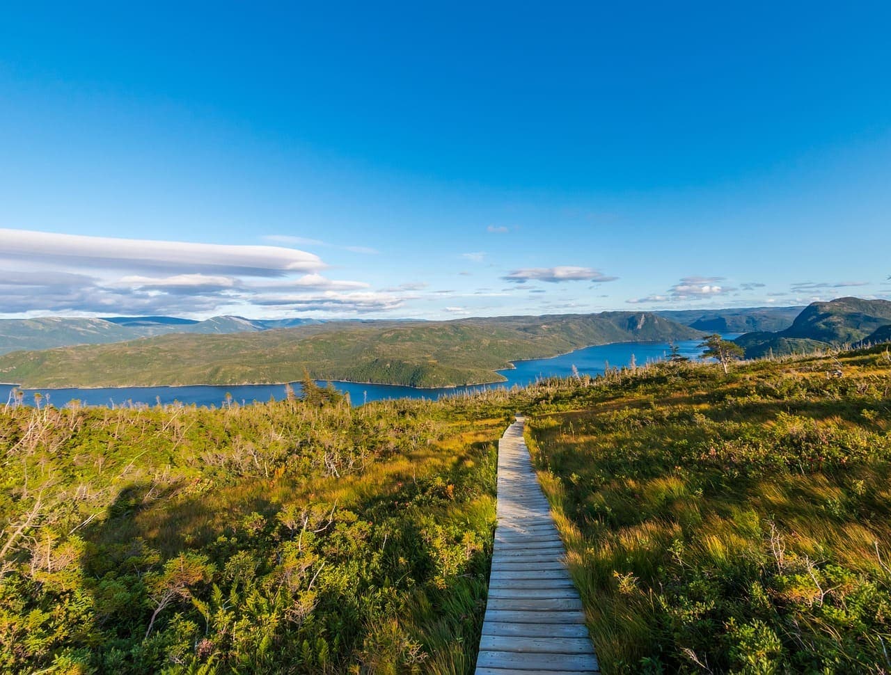 Wooden boardwalk trail running through a grassy landscape with lakes and hills visible in the distance.