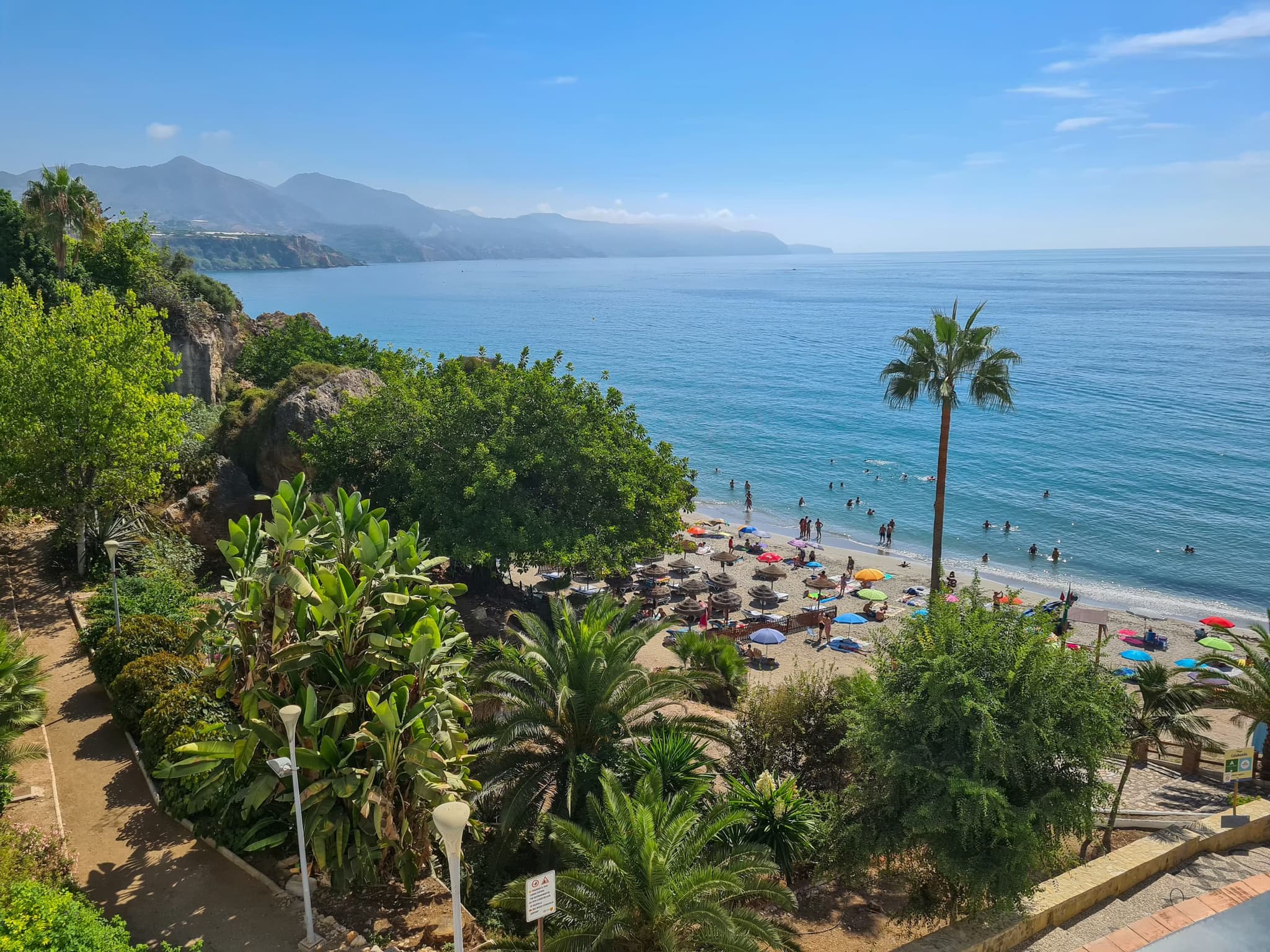 A scenic beach with colorful umbrellas, palm trees, and clear blue water. People swim and relax under a bright sky, framed by distant mountains.