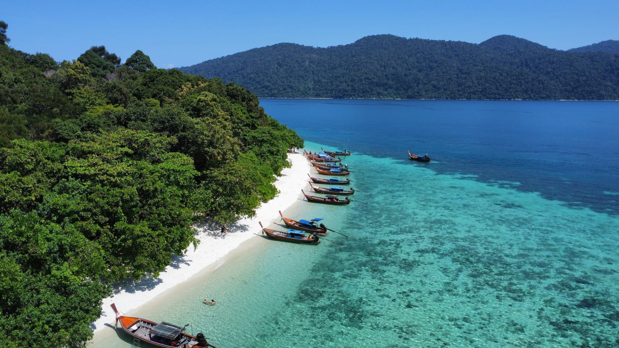 Boats lined up on the white sand in koh lipe thailand surrounded by clear turqoise water and lush green trees and mountains