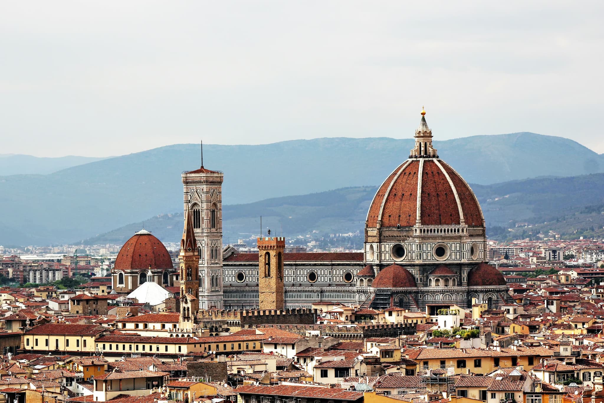 Panoramic view of Florence with the iconic Florence Cathedral, featuring its massive red-brick dome, and intricate bell tower, set against distant mountains.