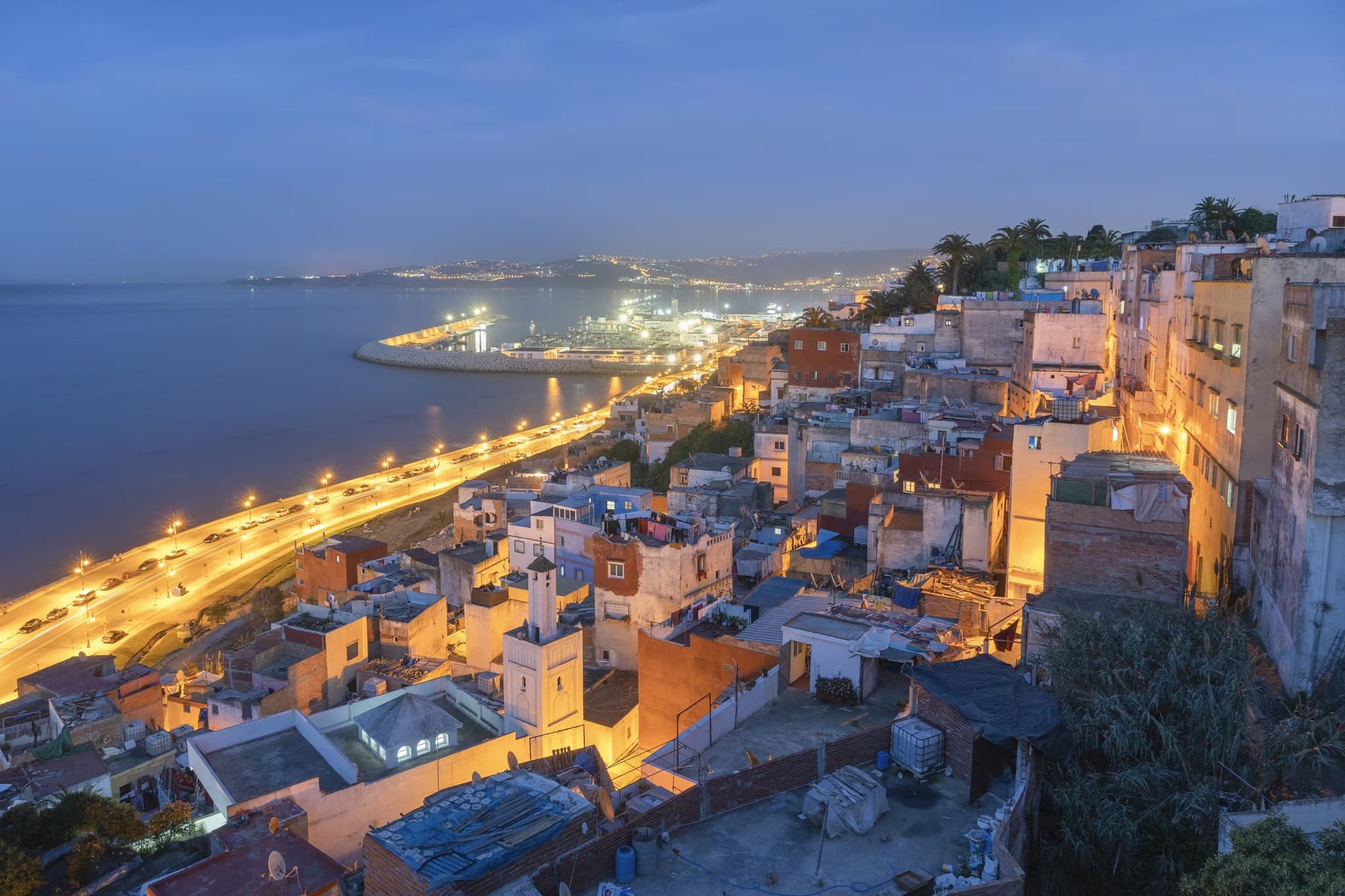 Hillside view of Tangier at dusk, featuring colorful buildings illuminated with streetlights and a curving seaside road beside a calm sea.