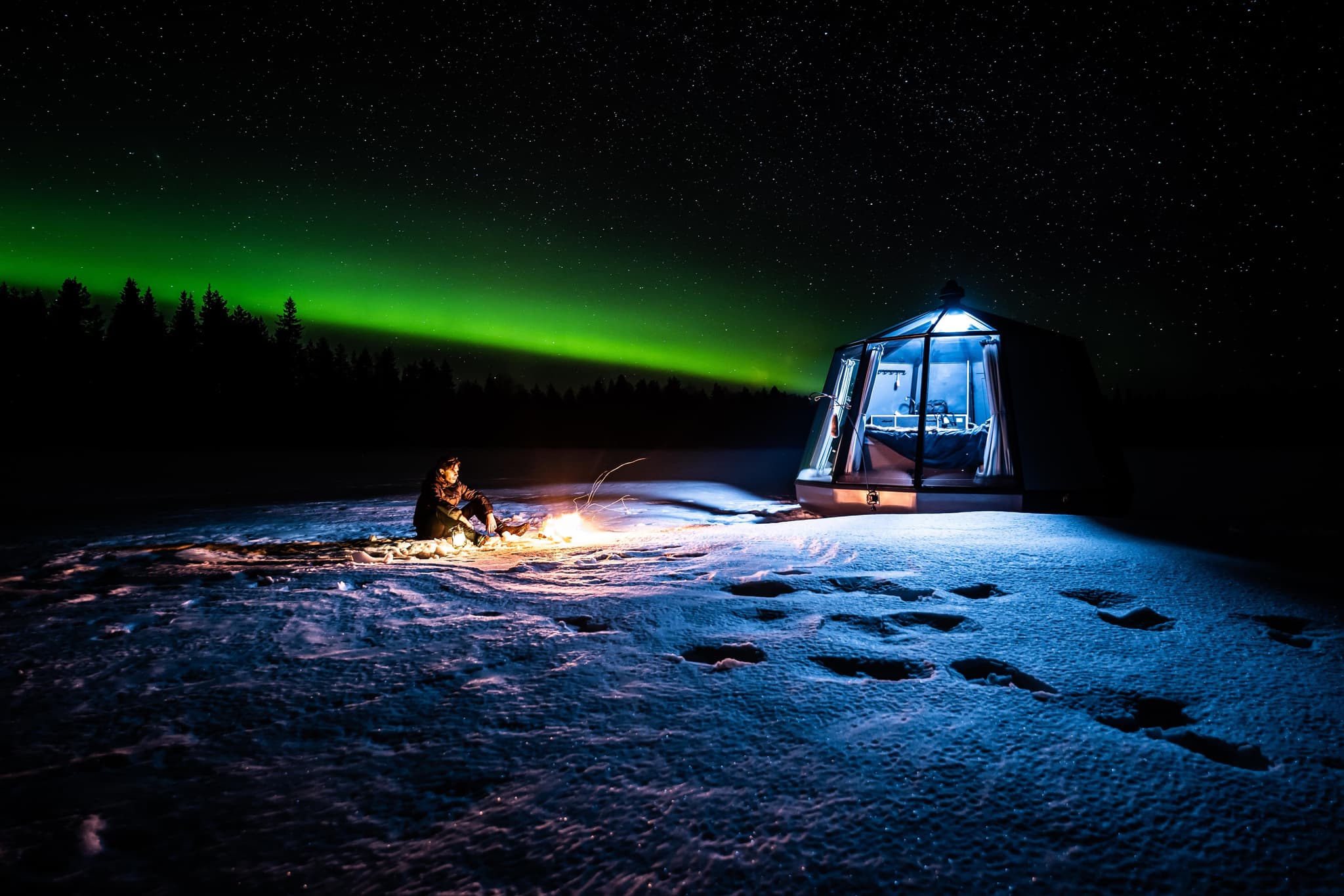 a person sitting next to a glass iglo at night when green aurora lights appear