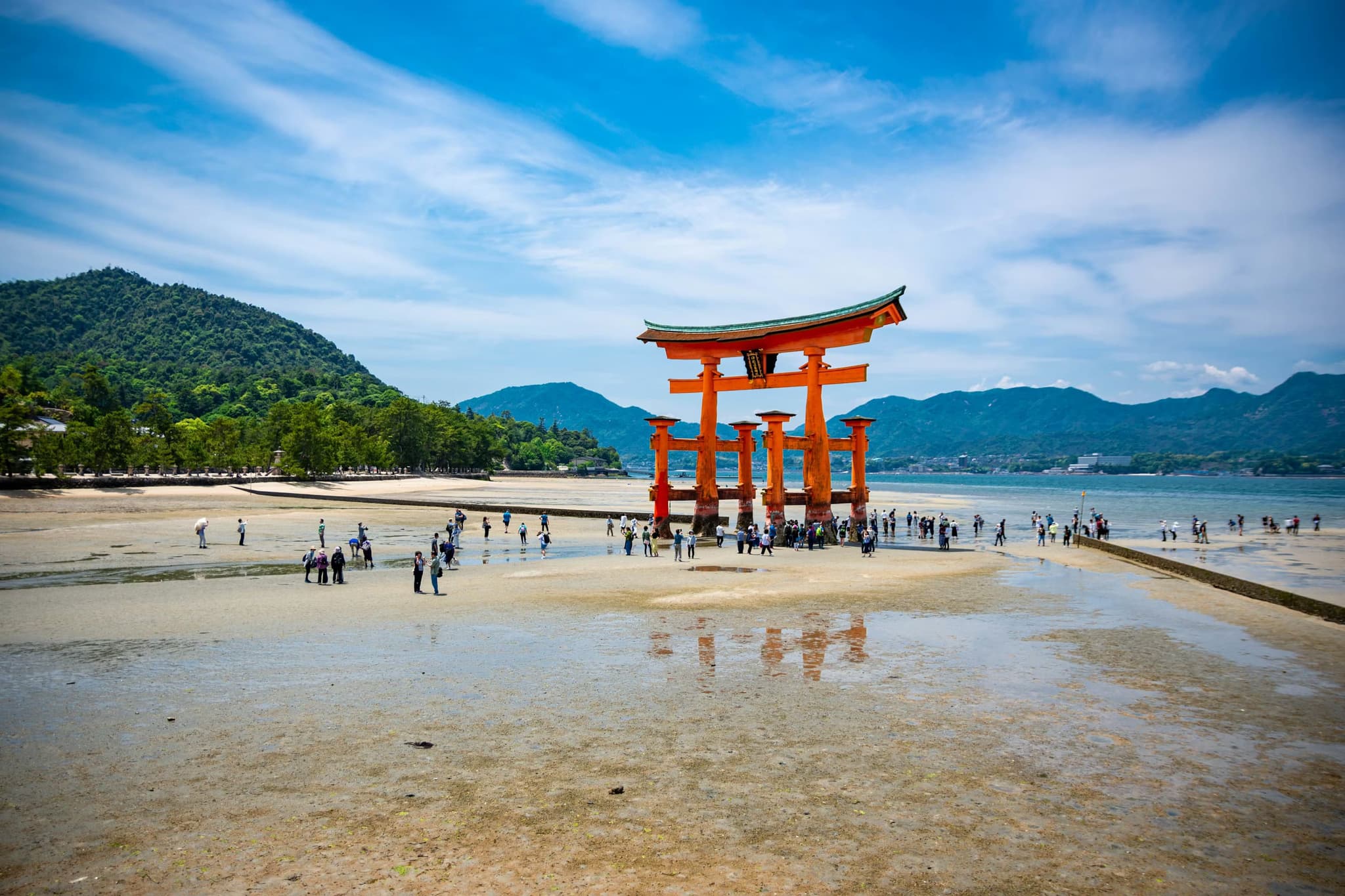 A bright red torii gate stands on wet sand surrounded by people, with lush green mountains and a vibrant blue sky in the background, evoking a serene atmosphere.