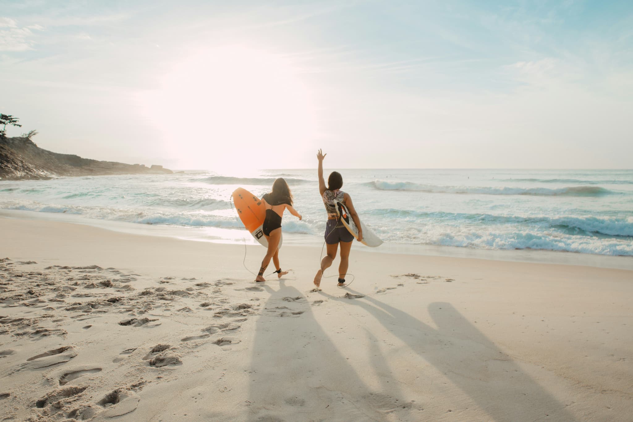 Two females walking with surf boards on the sand toward the sea