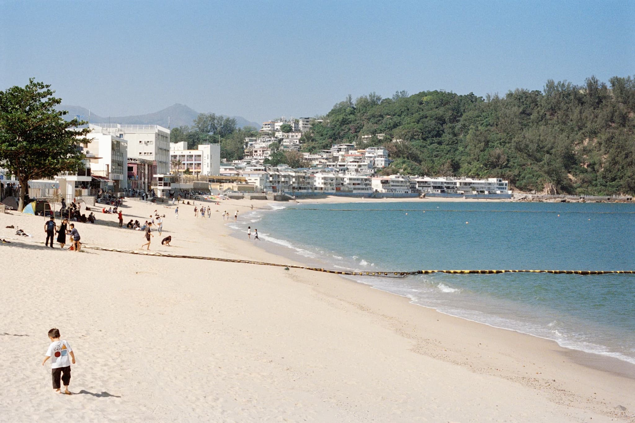 A sunny beach with soft, golden sand, a few people walking and relaxing. Clear blue water on the right meets green hills and white buildings.