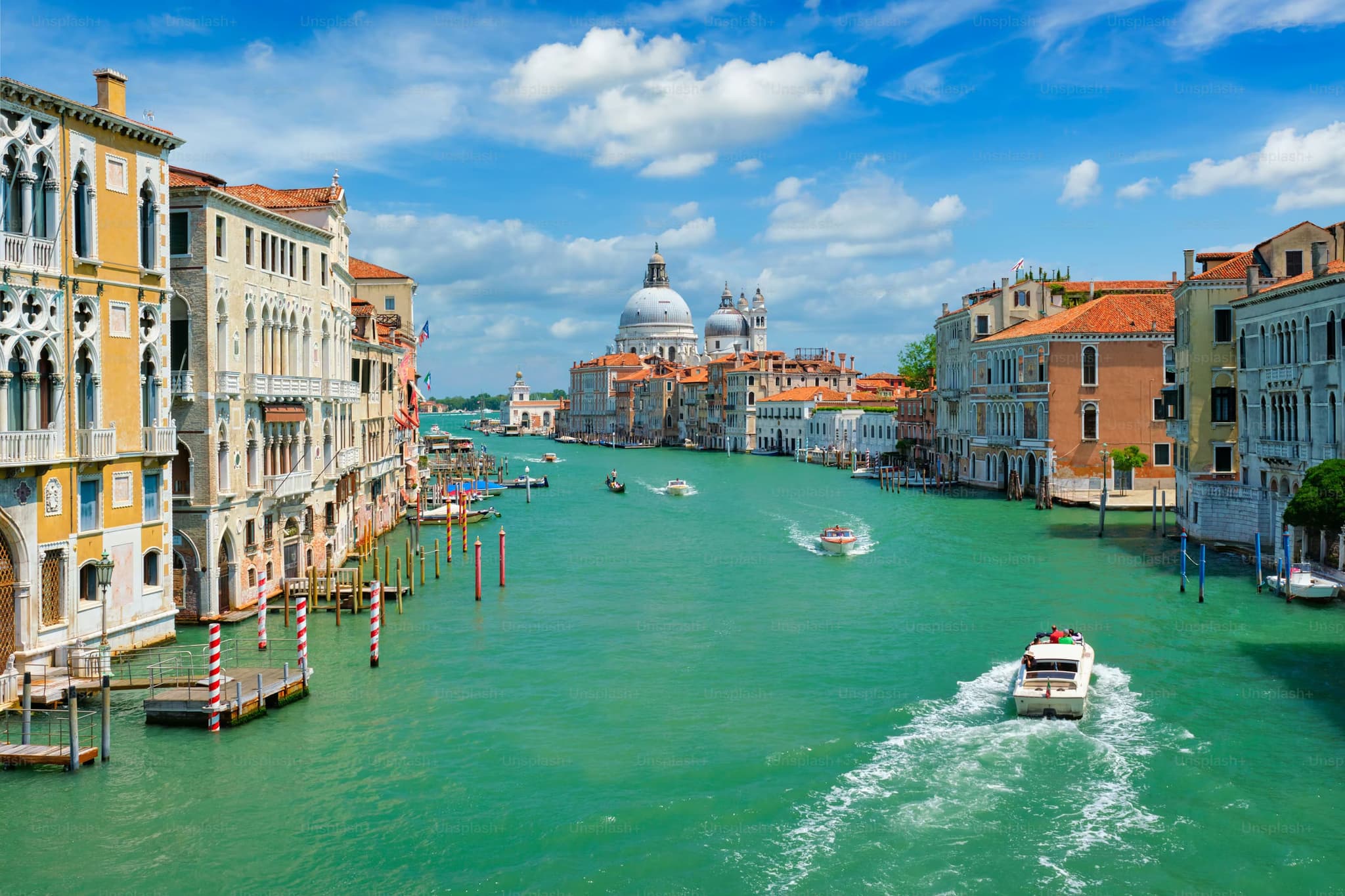 Venice's Grand Canal under a bright blue sky, with boats navigating the green water. Historic buildings flank the canal, leading towards a domed basilica.
