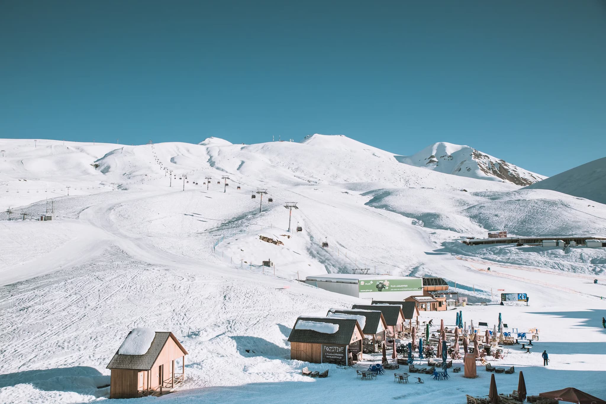 Snow-covered ski resort with wooden cabins and outdoor seating under clear blue skies. Ski lifts lead up powdery slopes, suggesting a serene winter getaway.