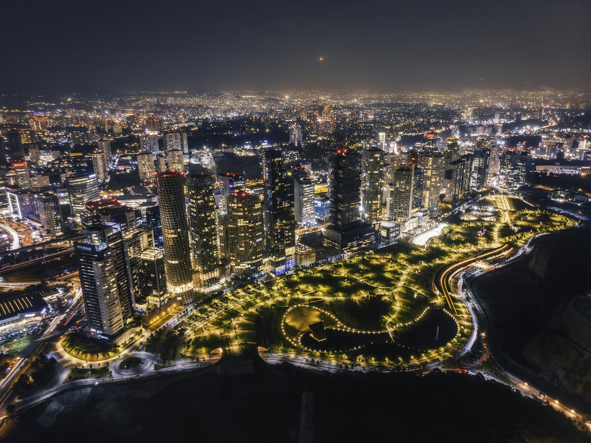 Aerial view of a city at night with skyscrapers illuminated, surrounded by a brightly lit park. The cityscape extends into the distance, creating a vibrant urban glow.