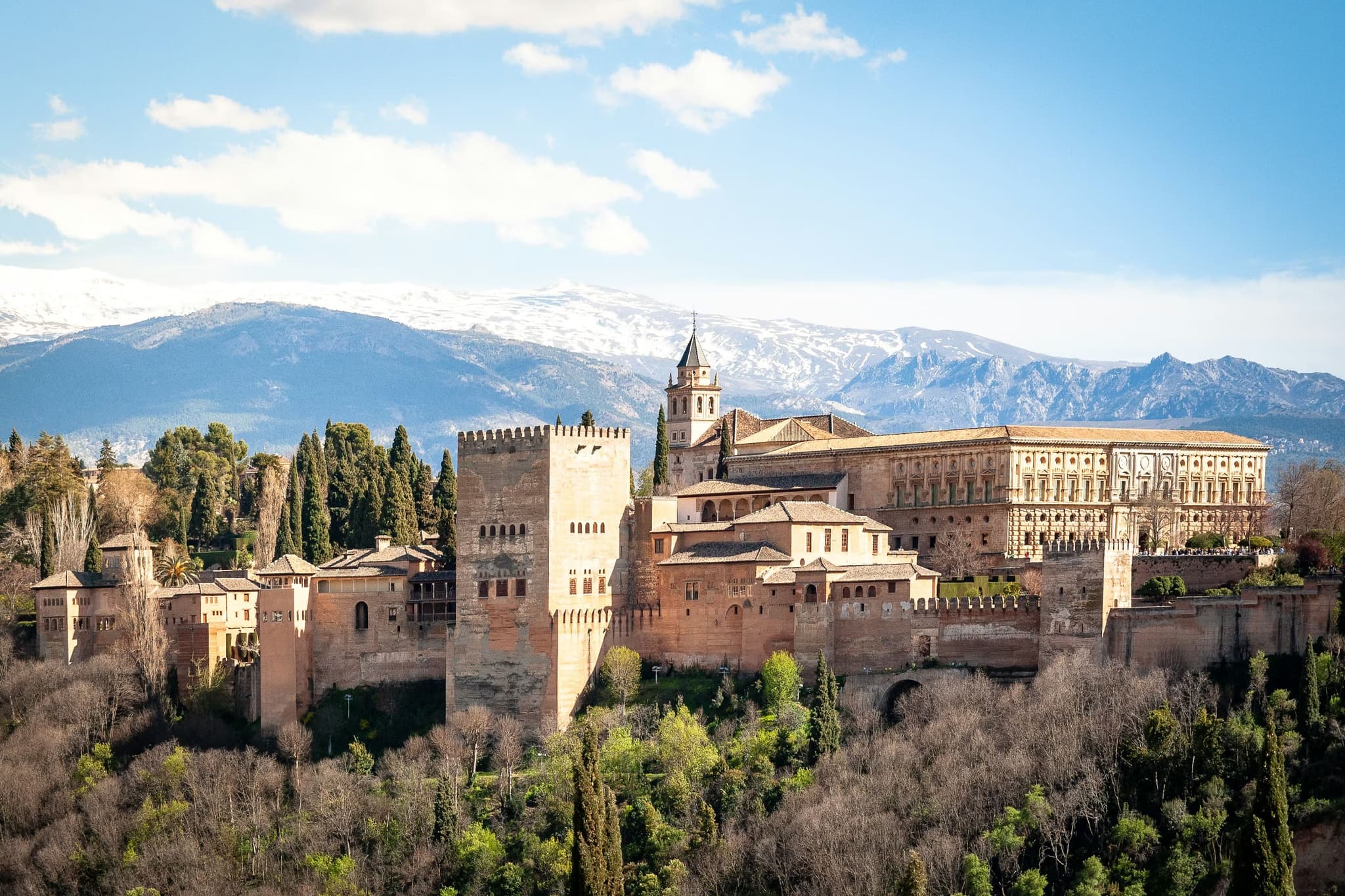 A sprawling fortress with tall, sandy-colored towers and walls surrounded by lush greenery, set against a clear blue sky and snow-capped mountains.