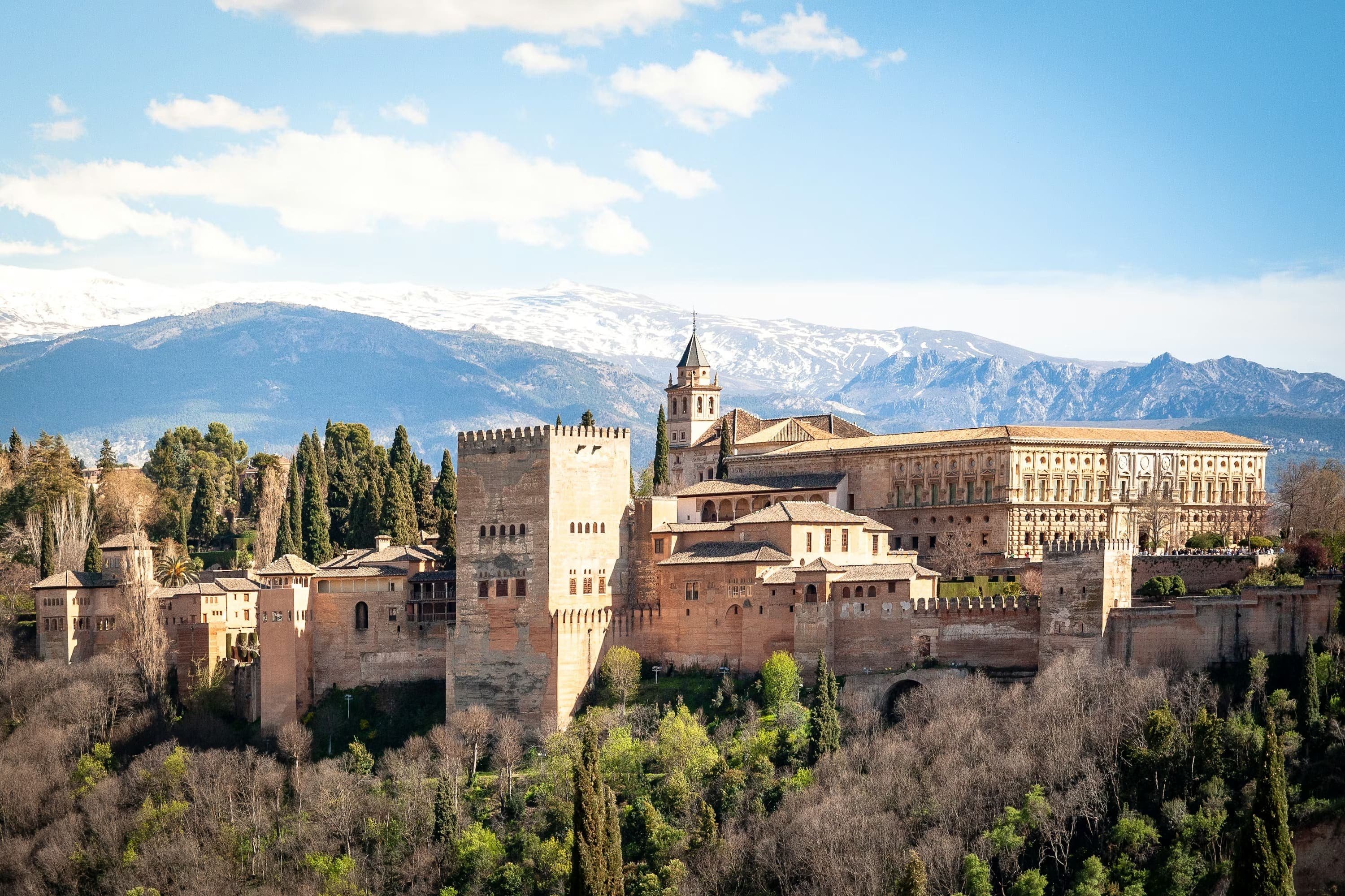 A sprawling fortress with tall, sandy-colored towers and walls surrounded by lush greenery, set against a clear blue sky and snow-capped mountains.