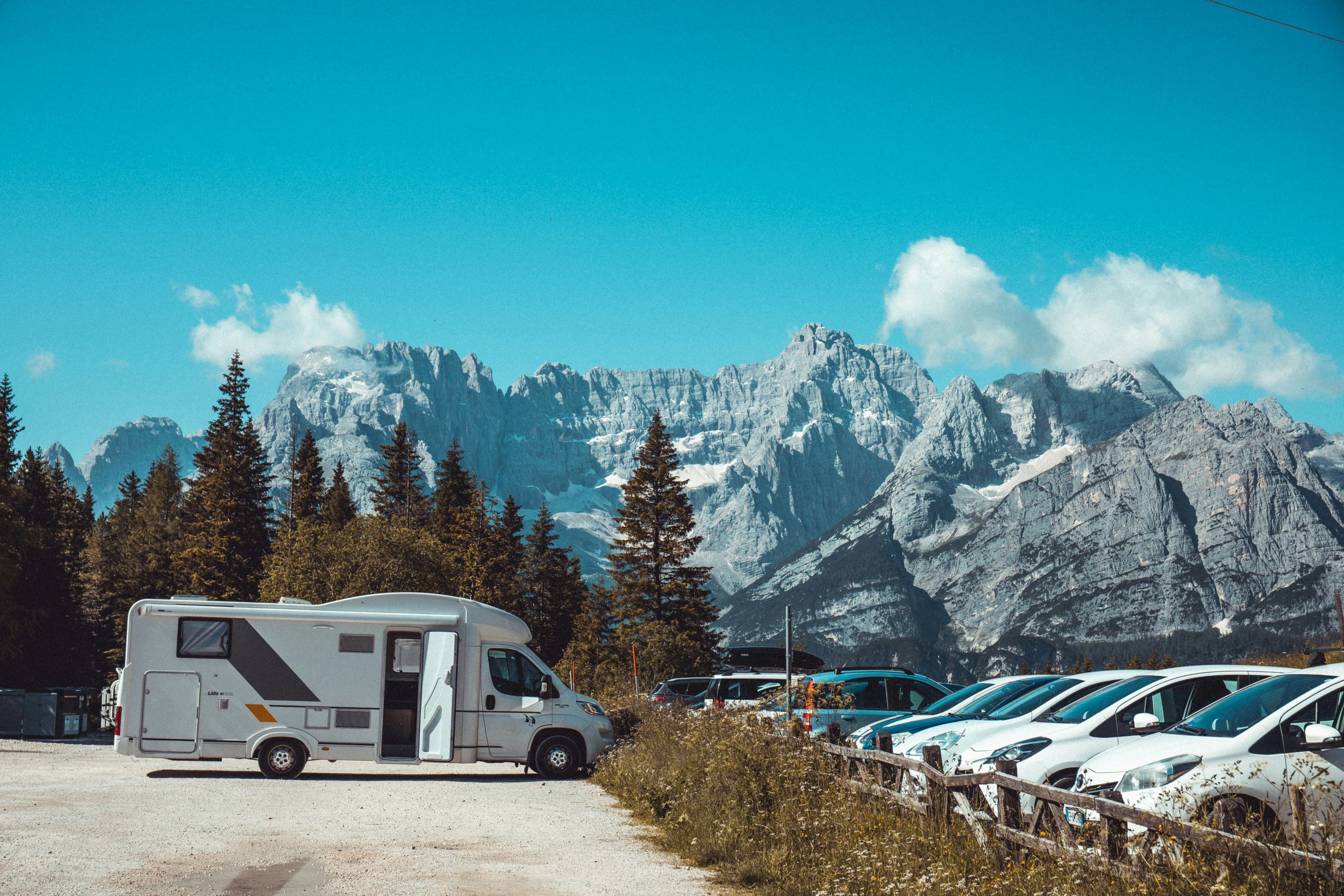 Campervan in front of a mountain range with snowy mountains, and a parking lot with campers on the right