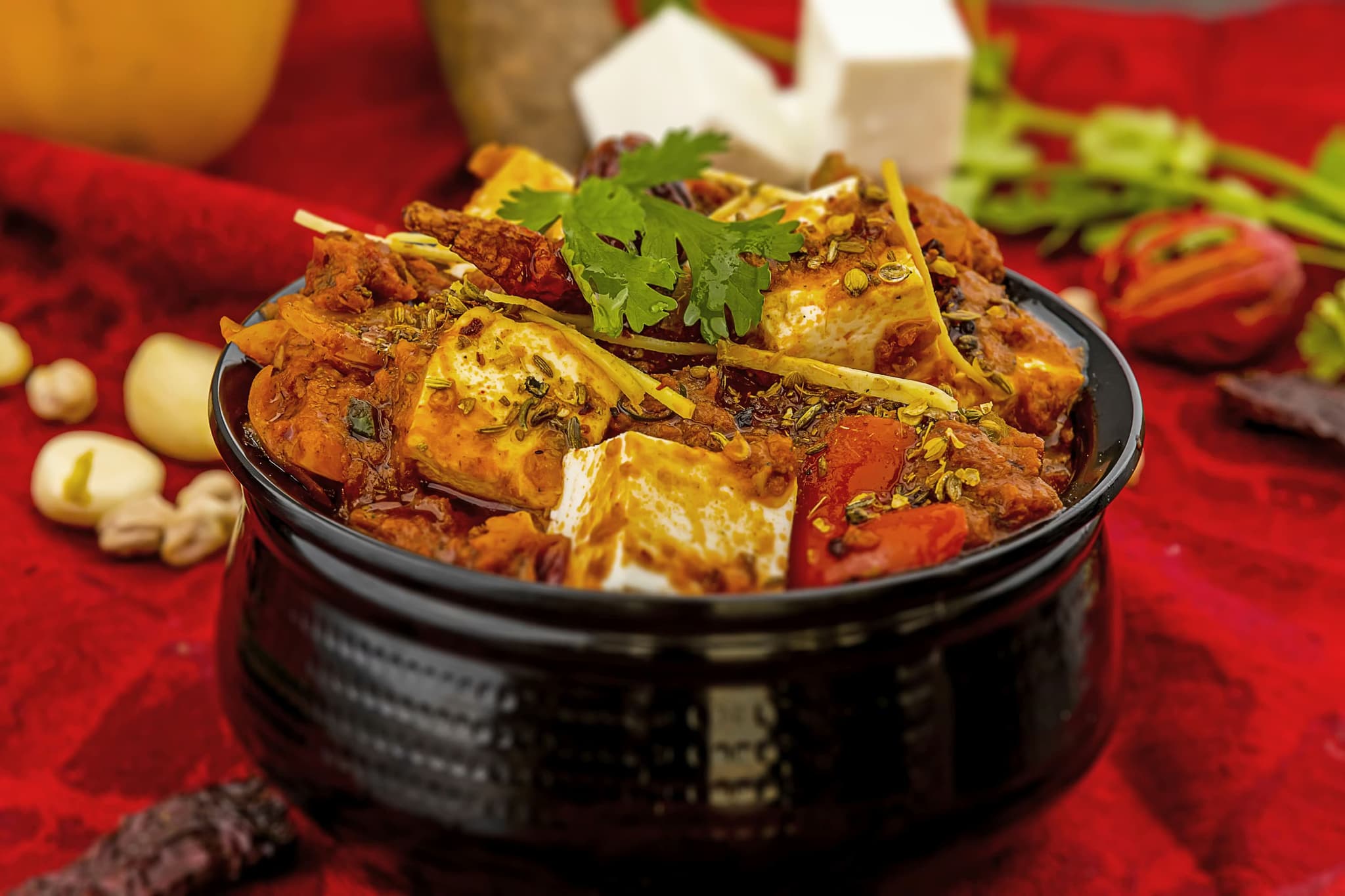 A black bowl filled with vibrant paneer curry, garnished with cilantro and spices, set on a rich red cloth with visible garlic and herbs nearby.