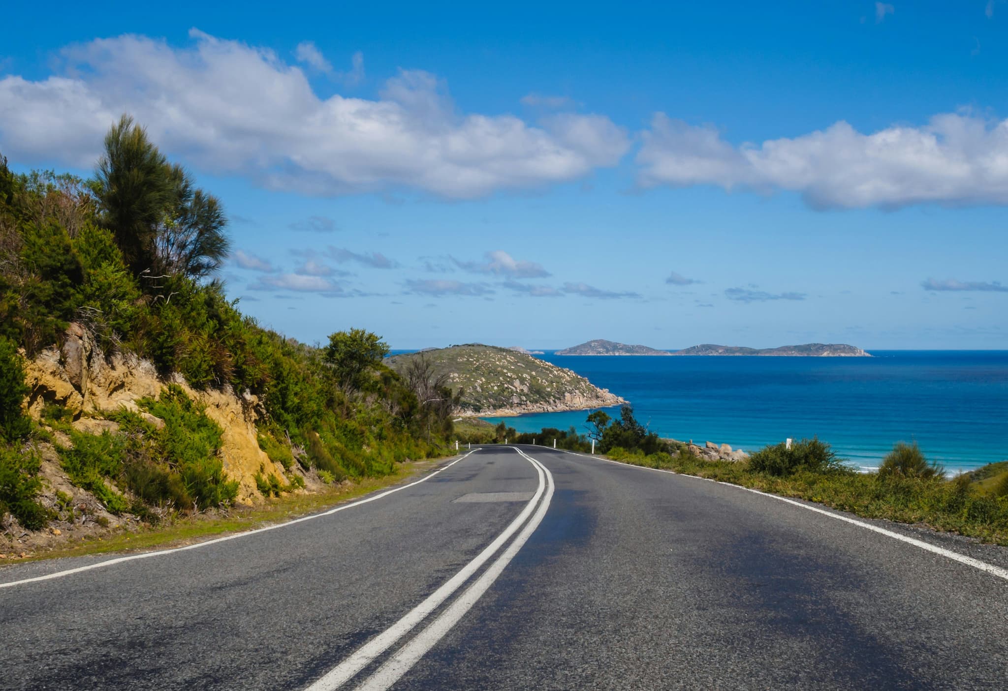 Windy road with two lanes in australia surrounded by hills, and turqoise beach water, clouds visible in the sky