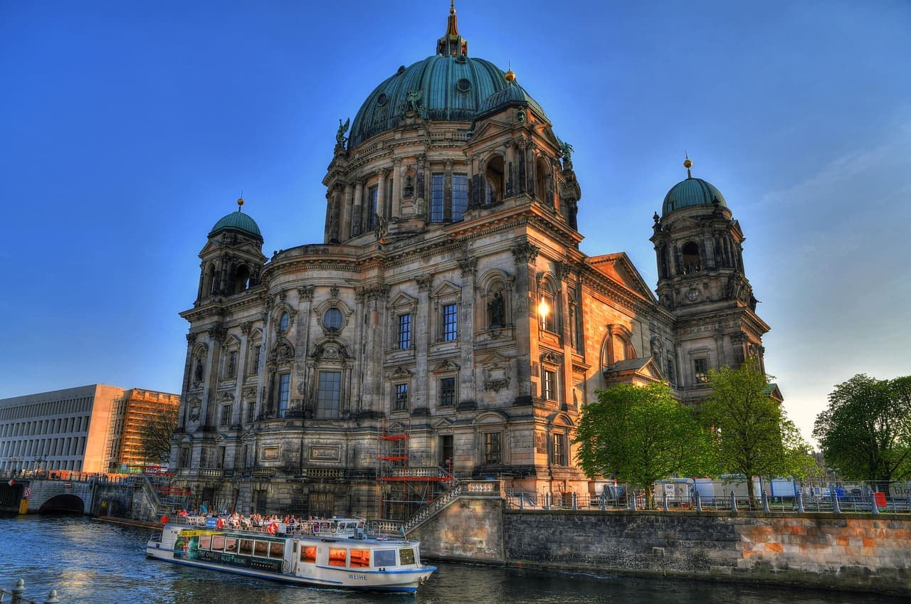 Berlin Cathedral at sunset with a river boat passing in front and historic buildings along the waterfront.