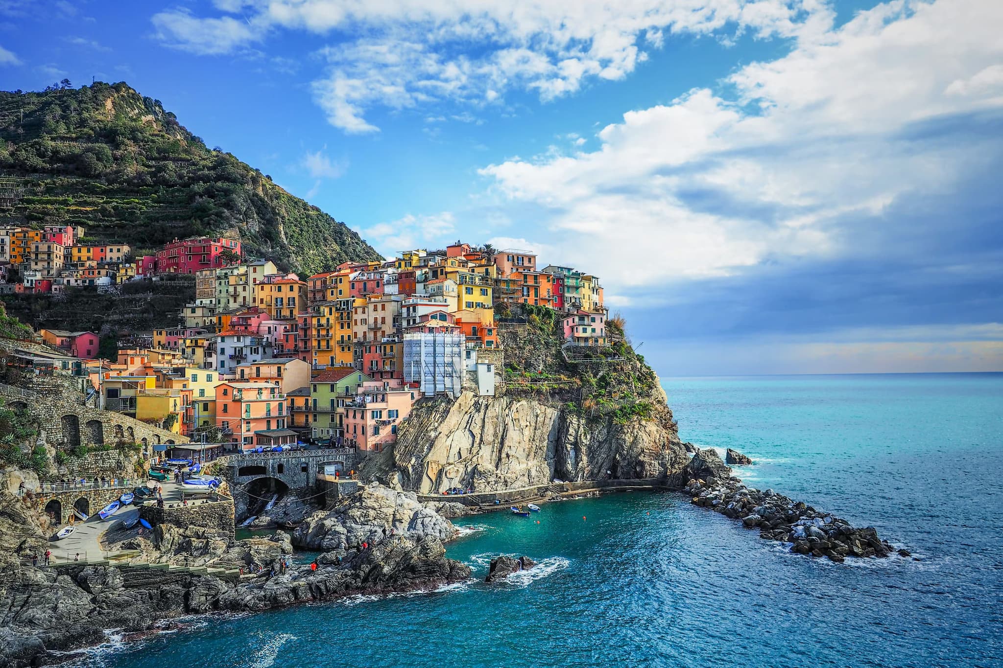 Colorful houses perched on a rocky cliff overlooking the turquoise Mediterranean Sea with a blue sky and scattered clouds above, evoking a serene coastal charm.