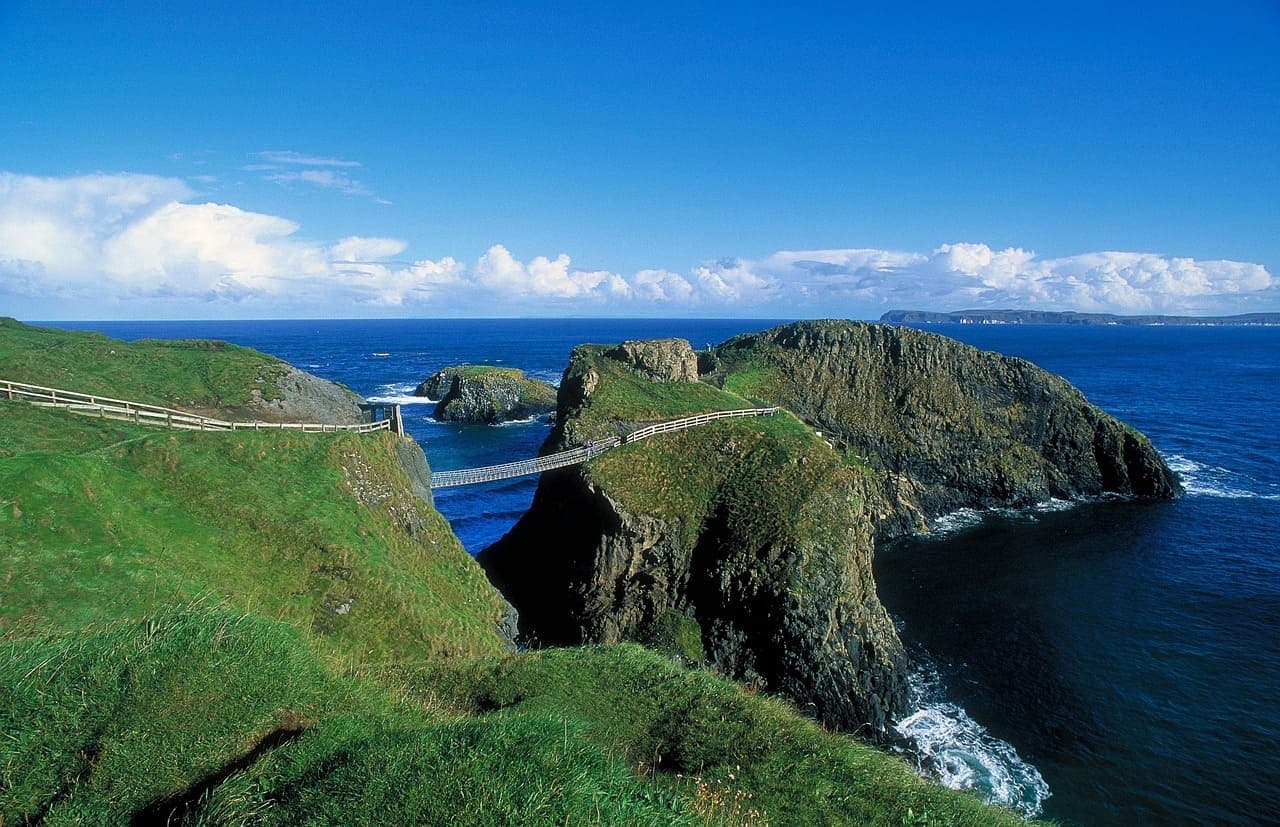 Cliffside coastal view in Northern Ireland featuring green grass, steep rock formations and the Carrick-a-Rede Rope Bridge stretching across the water.