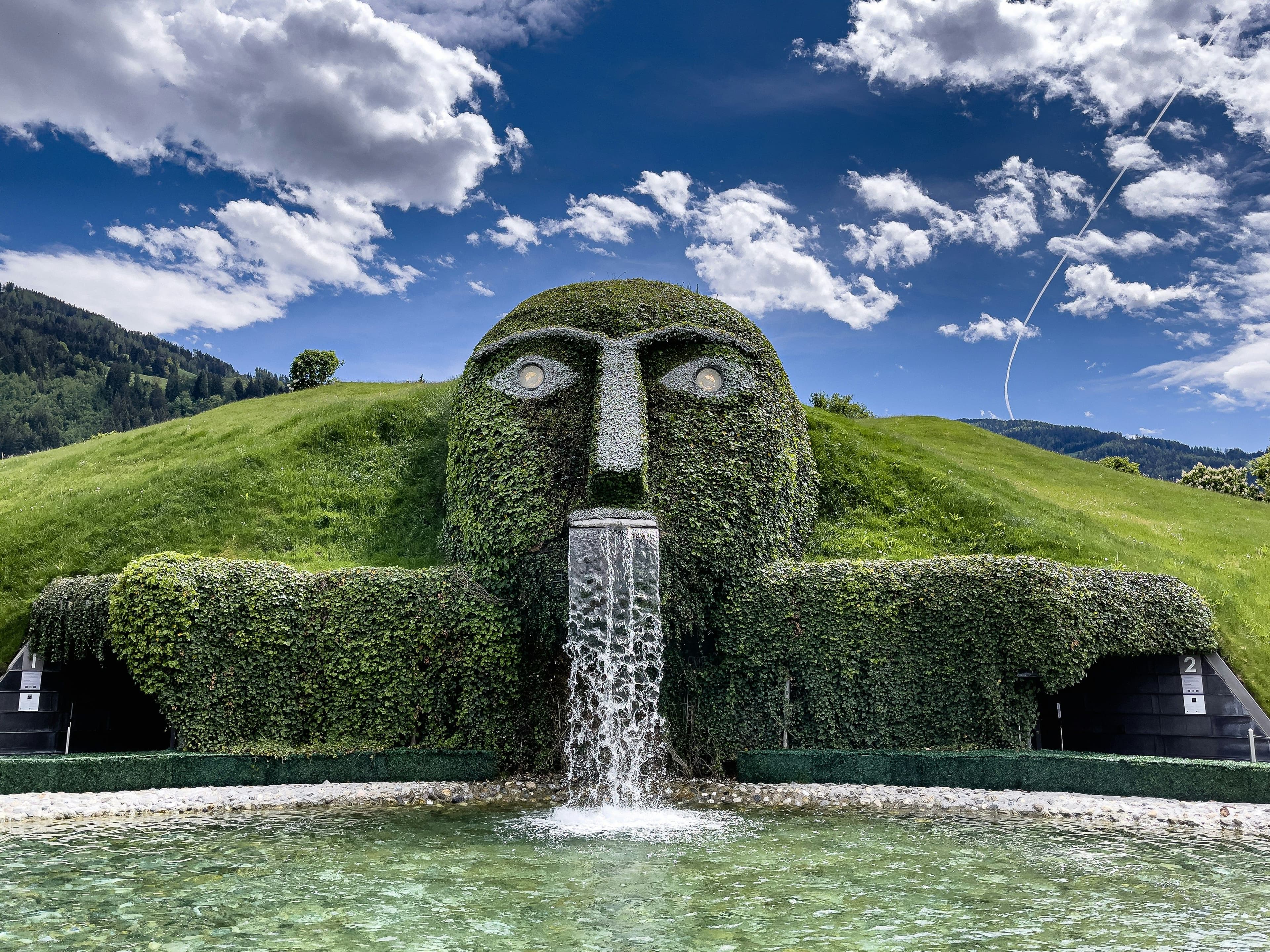 A landscape hill formed into the shape of a head with water from a waterfall coming out of its mouth into a fountain
