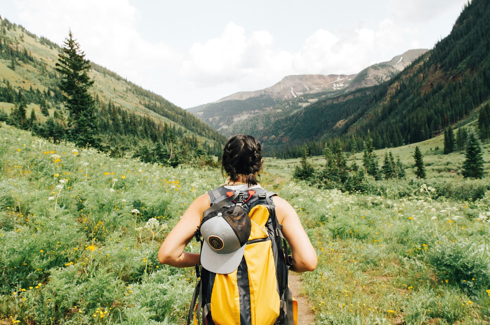 A picture from the back of a woman with yellow backback hiking in mountain green area