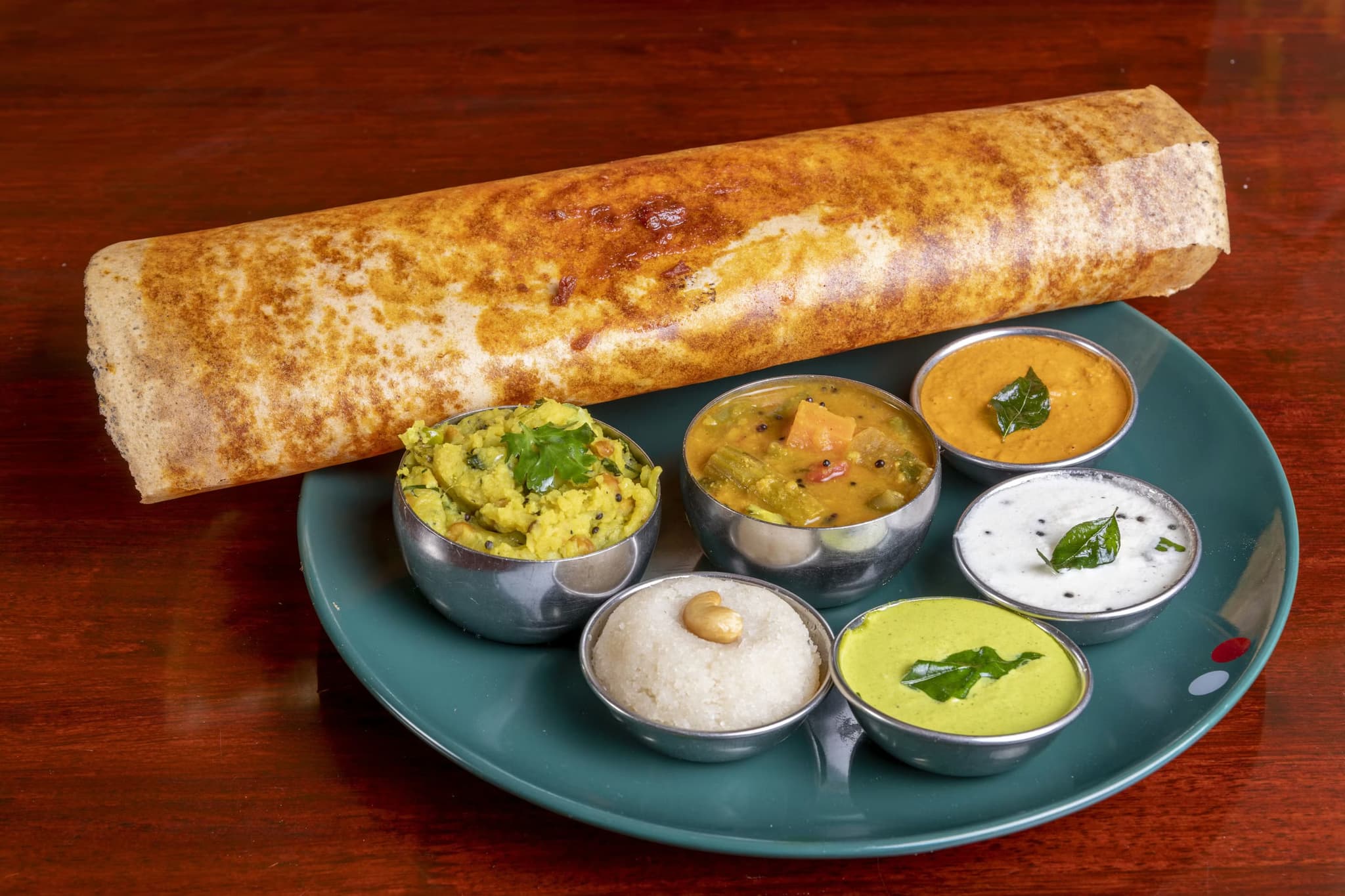 A crispy, golden-brown dosa is served on a green plate with six small metal bowls containing various colorful Indian chutneys and sambar, creating a vibrant and appetizing presentation.
