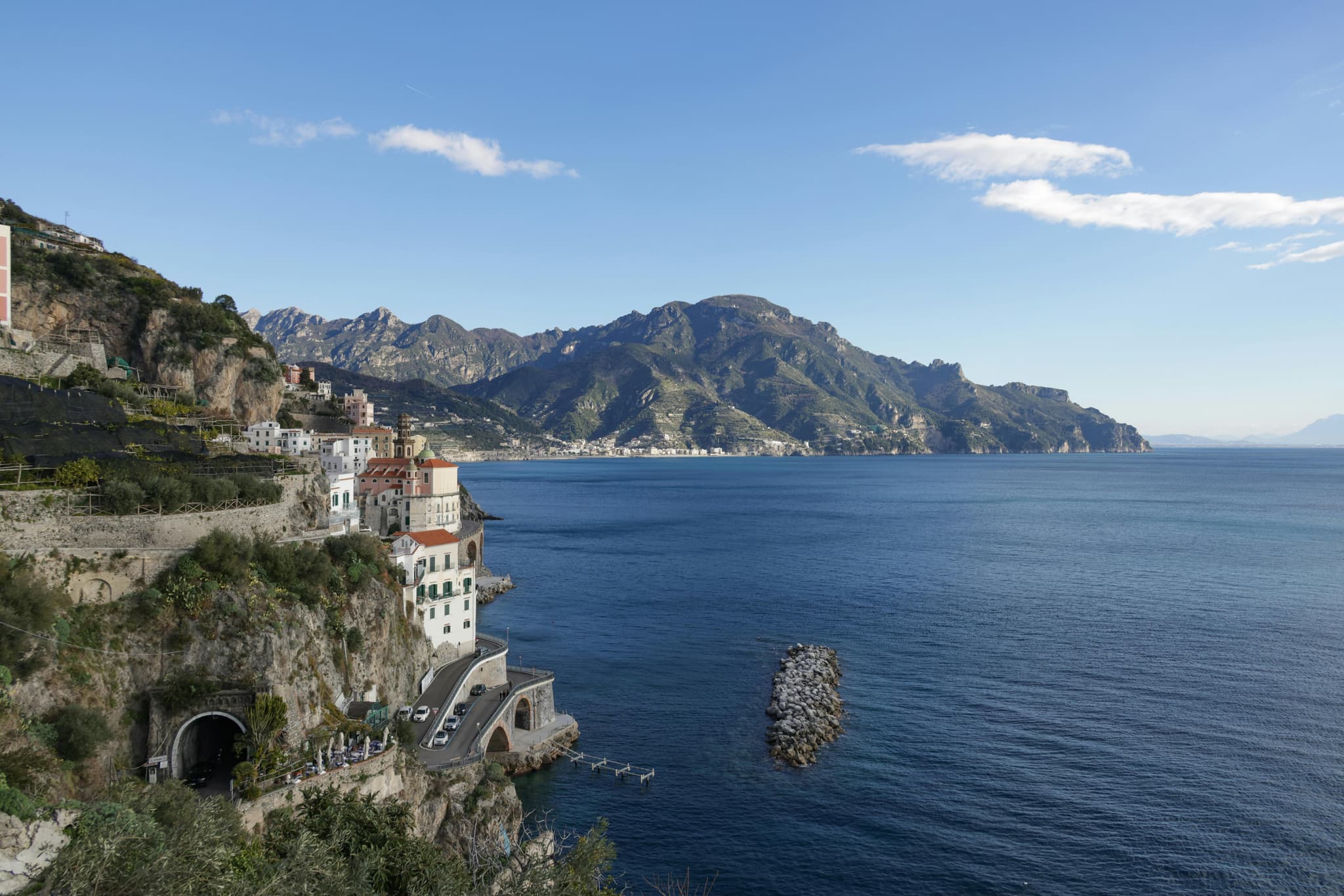 Coastal view of a serene blue sea with a mountainous backdrop. Cliffside houses and a winding road with a tunnel overlook the calm waters. Clear sky.