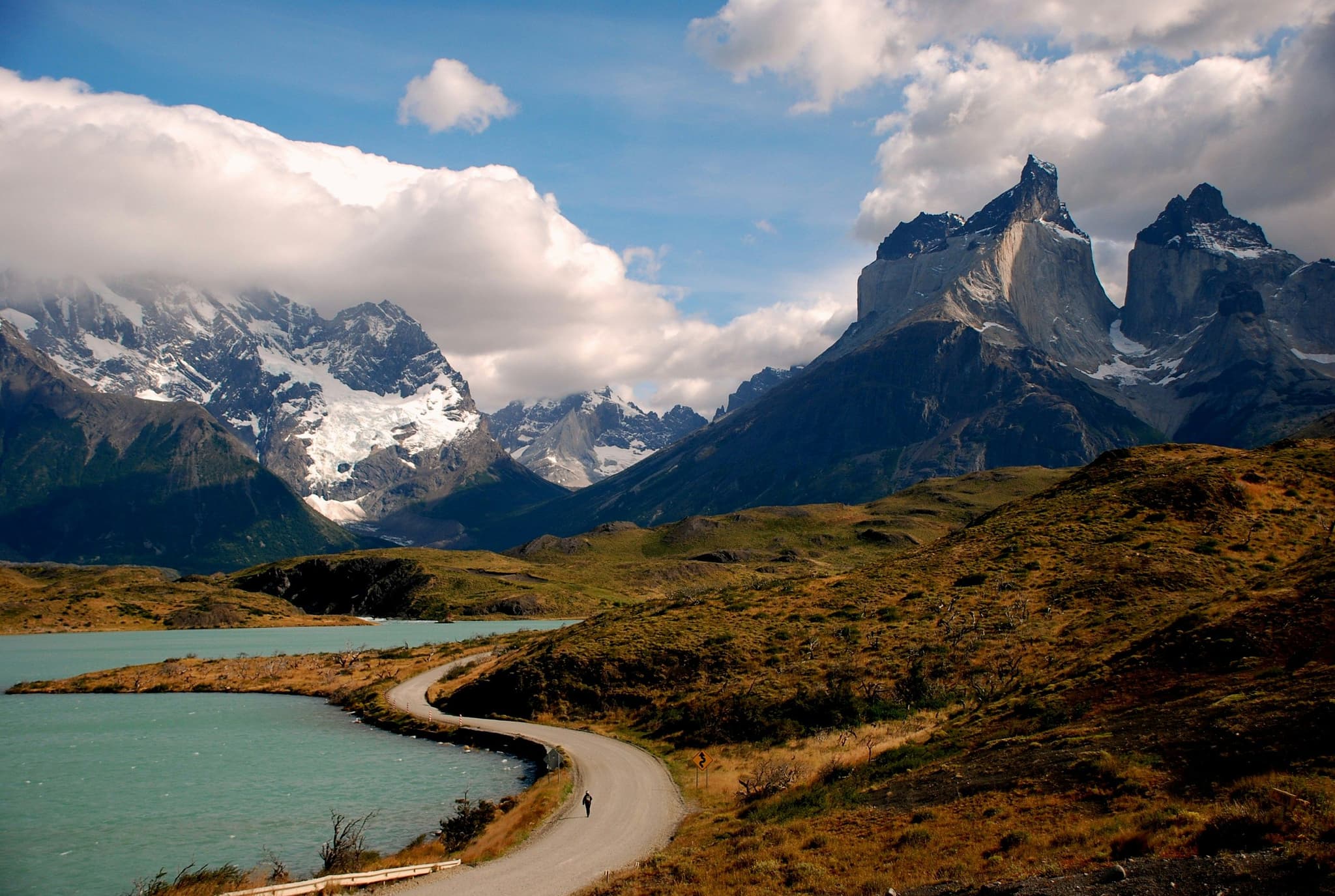 Windy road, blue lake and green grass surroundes by big rocky mountains and clouds