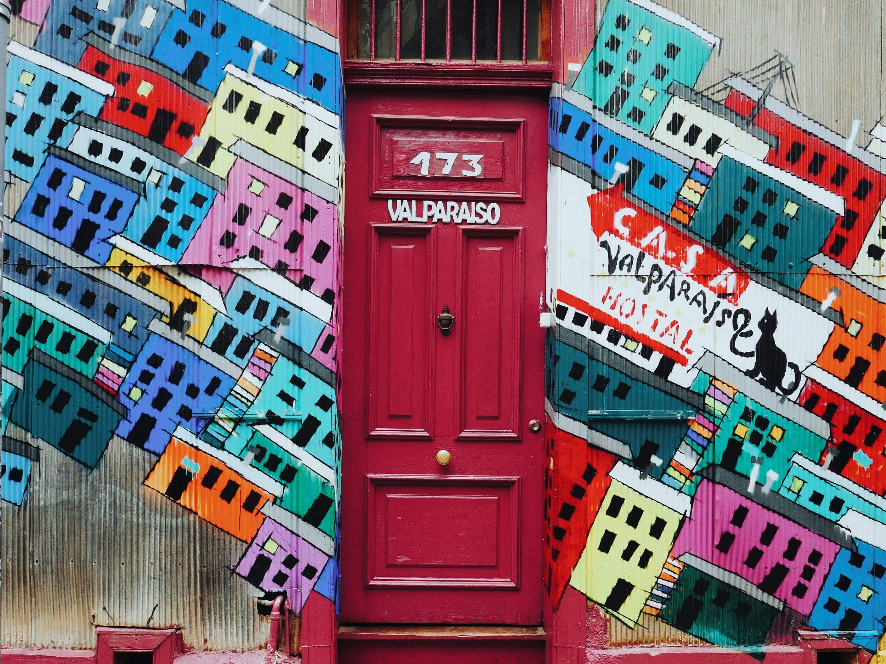 A red door with graffiti behind it of colorful houses of Valparaíso
