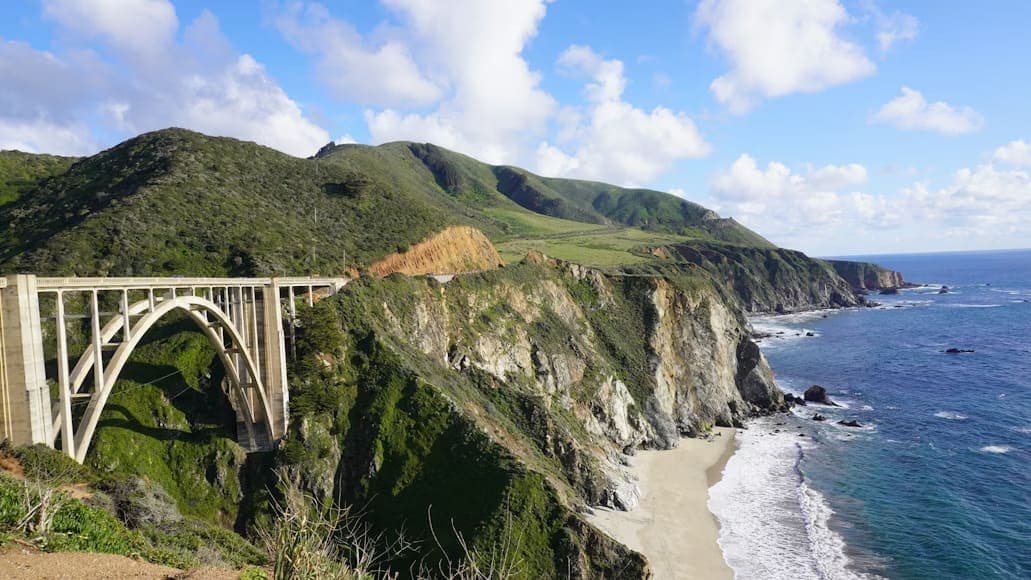 A sweeping coastal view features an arched concrete bridge spanning rugged cliffs, with lush green hills and a serene blue ocean under a partly cloudy sky.