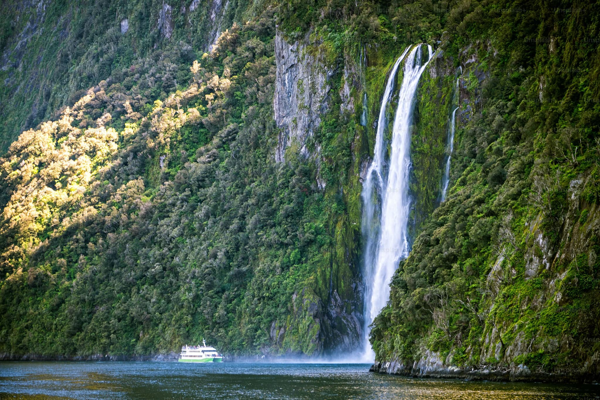 A majestic waterfall cascades down a lush, green cliff into a serene fjord. A small boat sails nearby, highlighting the scale and tranquility of the scene.