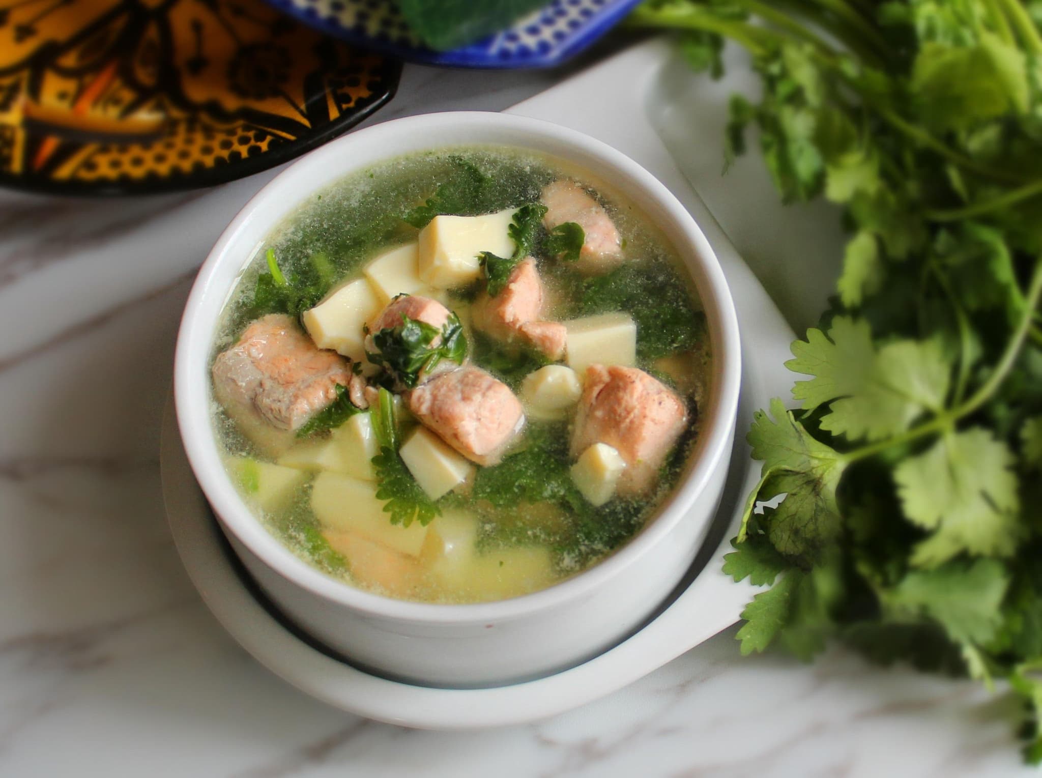 A bowl of soup with salmon inside on a table with coriander leaves