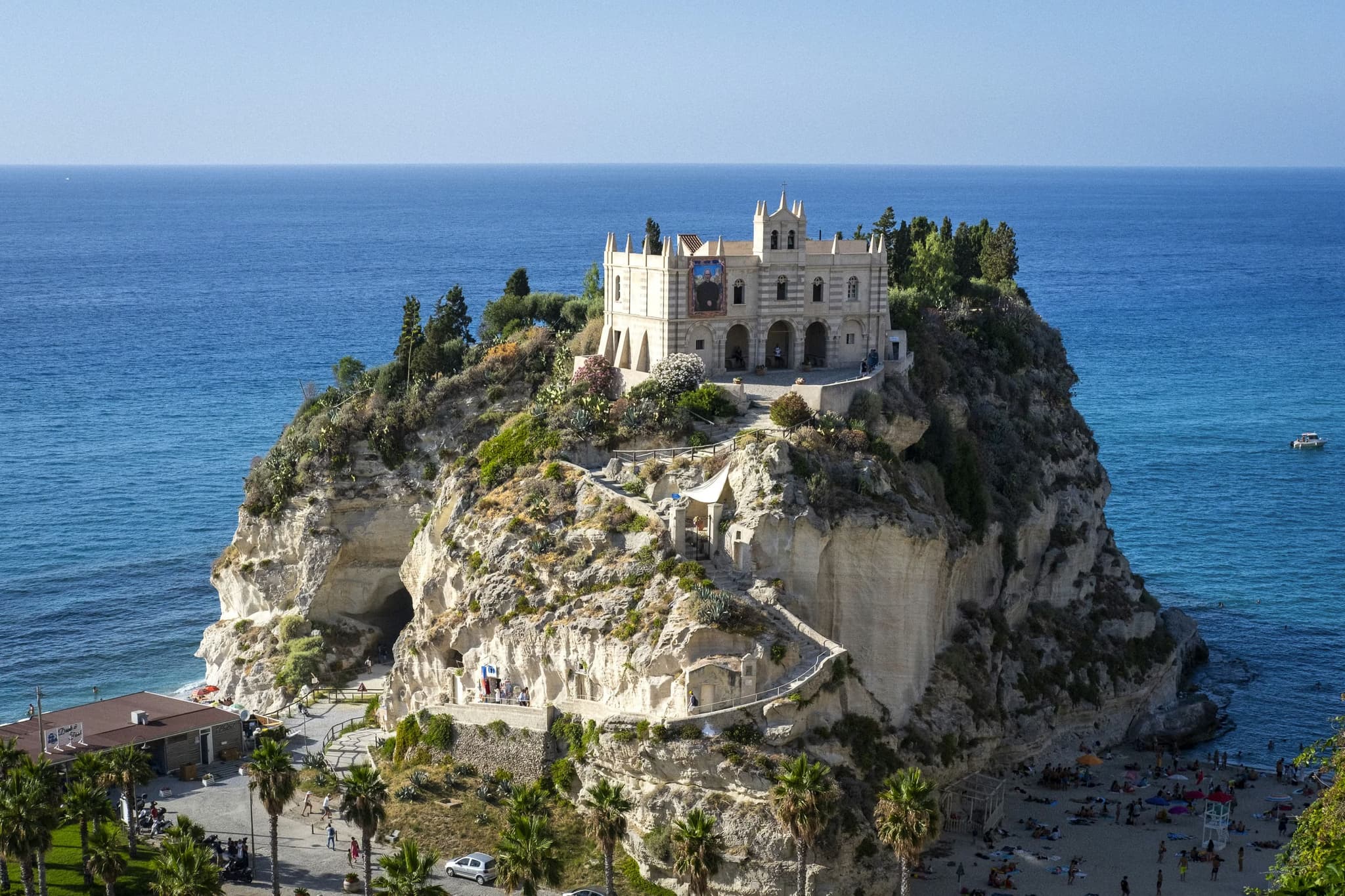 Historic monastery perched on a rocky cliff, overlooking the deep blue ocean. Lush greenery surrounds the structure, with a vibrant beach below.