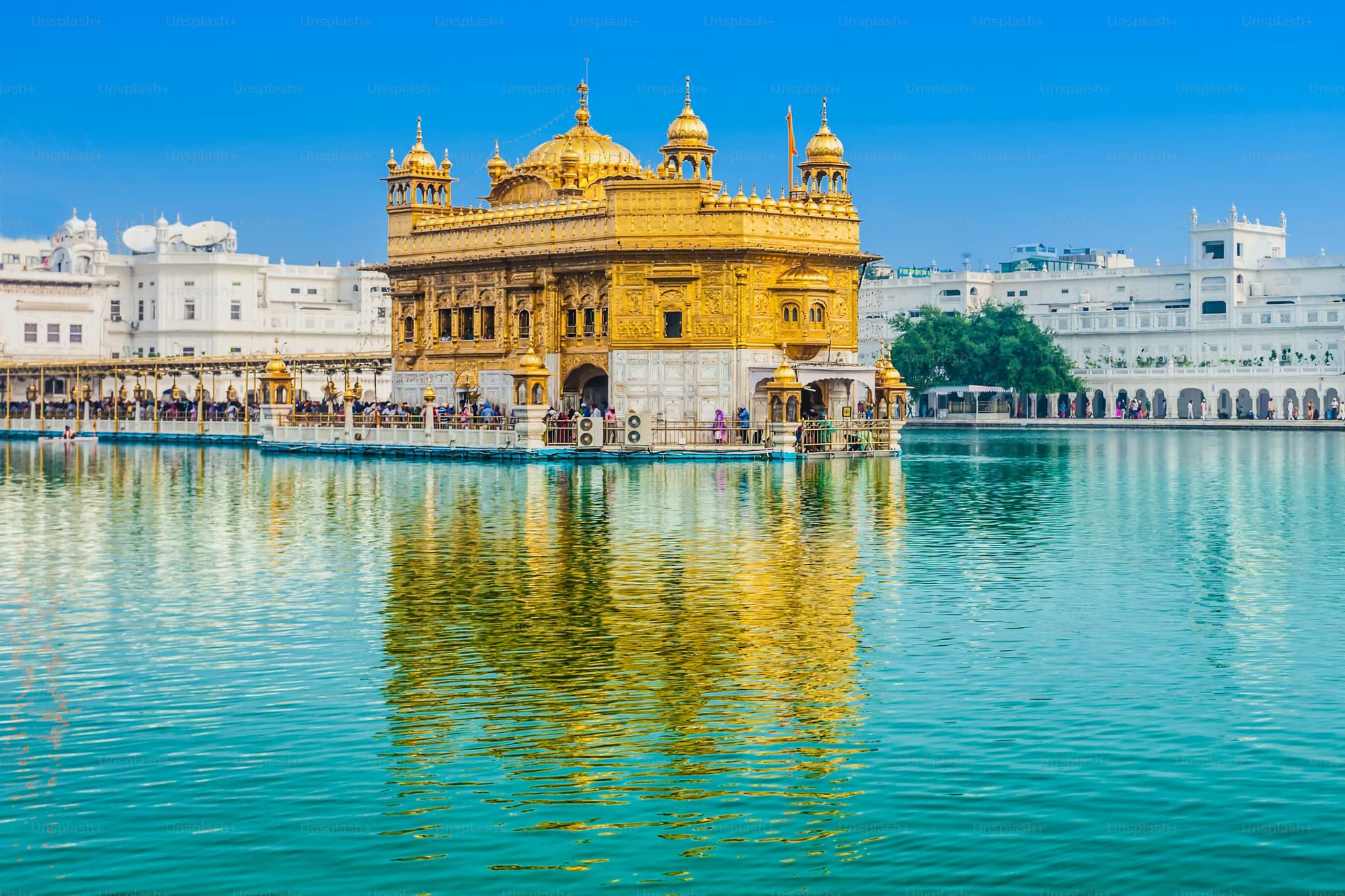 Golden Temple amidst blue water, reflecting its ornate gold architecture under a clear sky. Devotees surround the temple, creating a serene atmosphere.