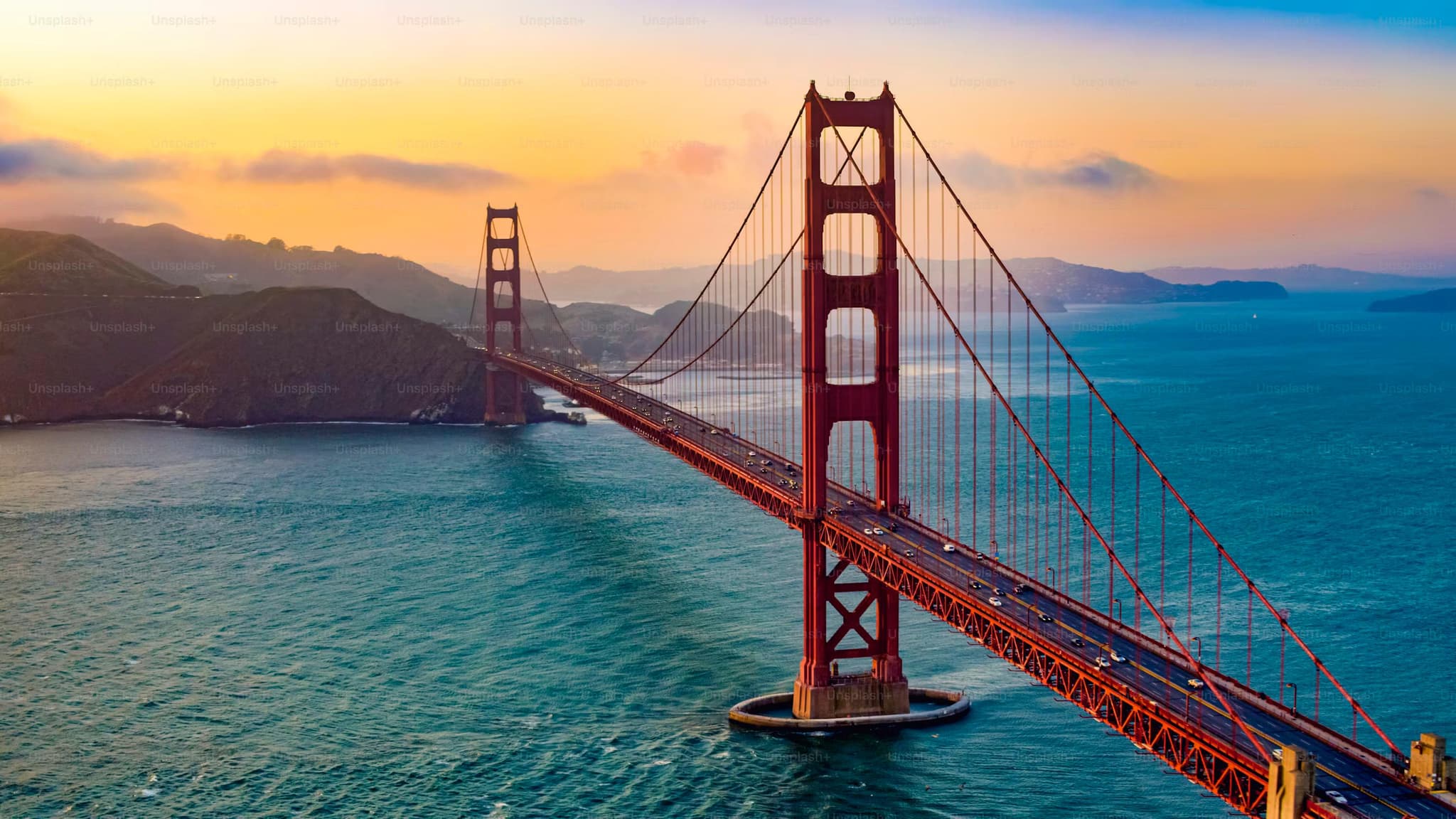 Curved bridge crossing rocky islands over calm ocean, with snowy mountains in the background under a cloud-dappled sky, conveying a serene mood.